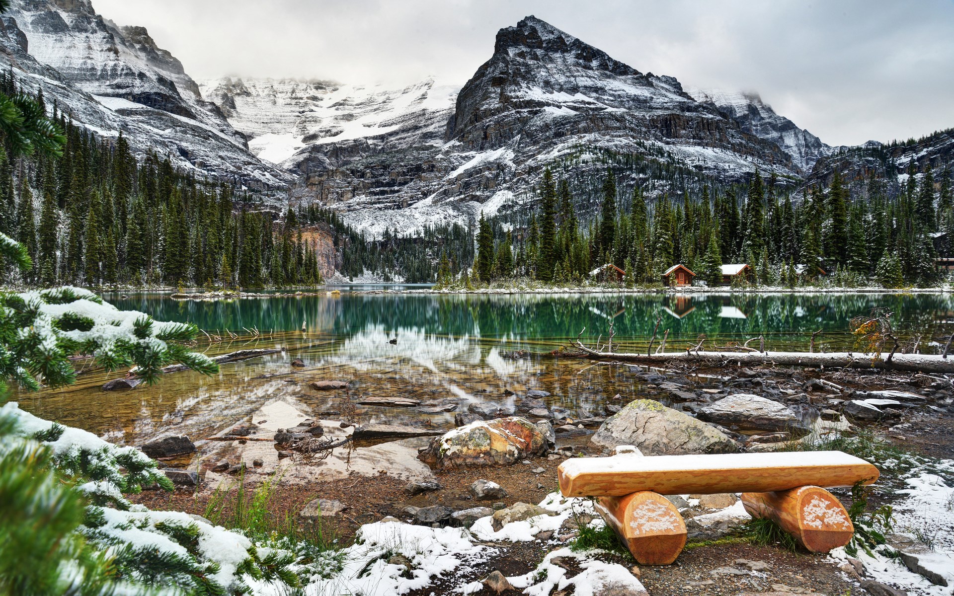 Bench by a snowy mountain lake with cabins HD Wallpaper