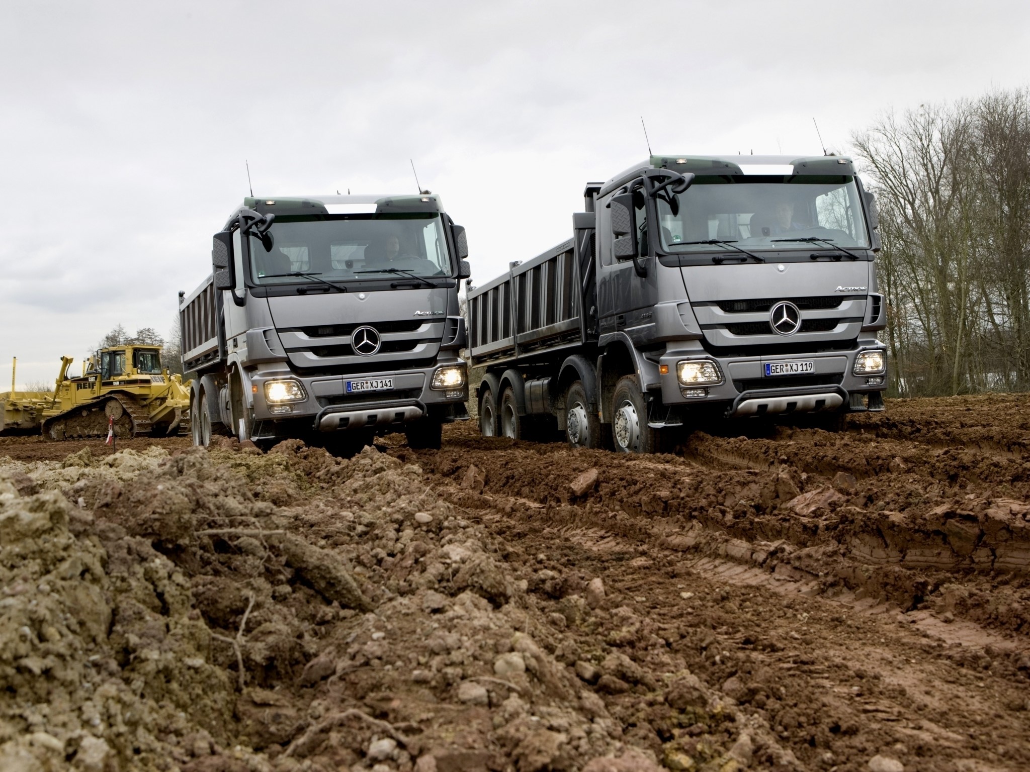 Two Mercedes-Benz trucks on a muddy construction site with a bulldozer HD Wallpaper