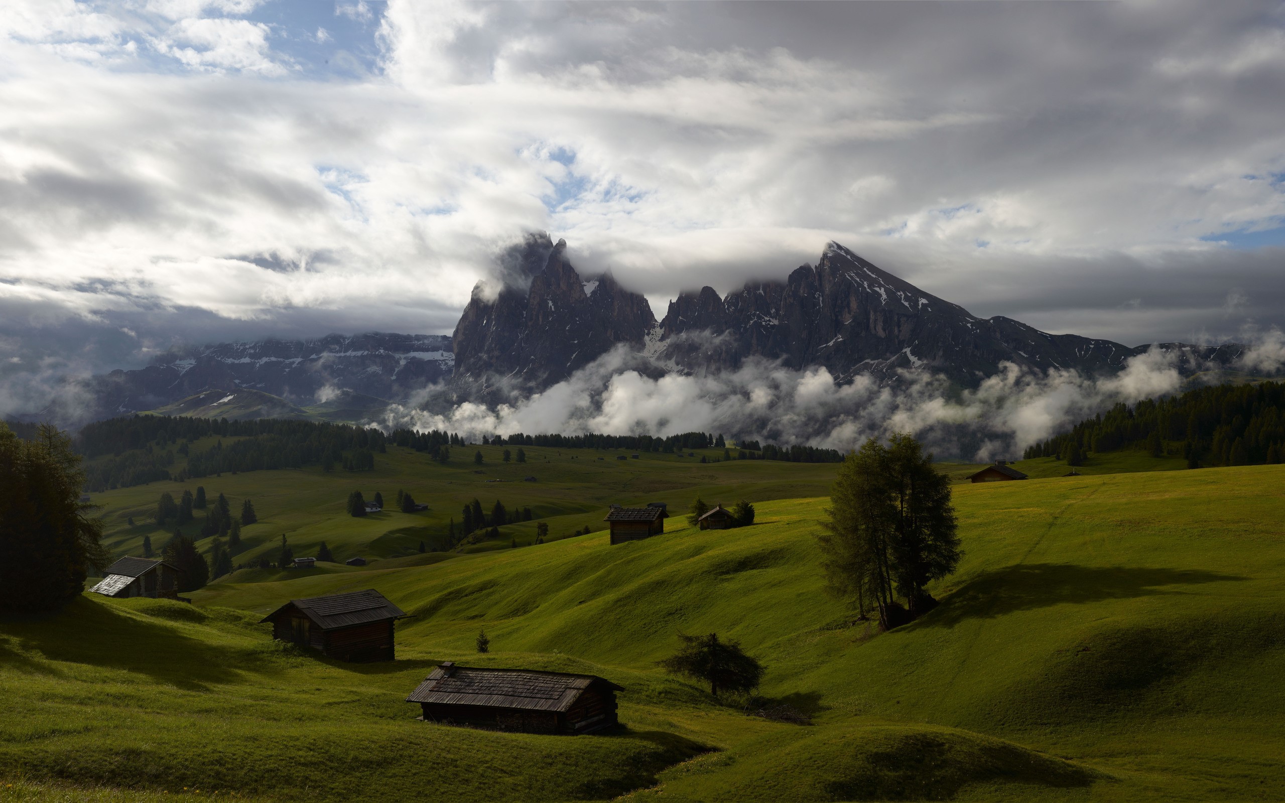 Mountains with clouds and small cabins in a valley HD Wallpaper