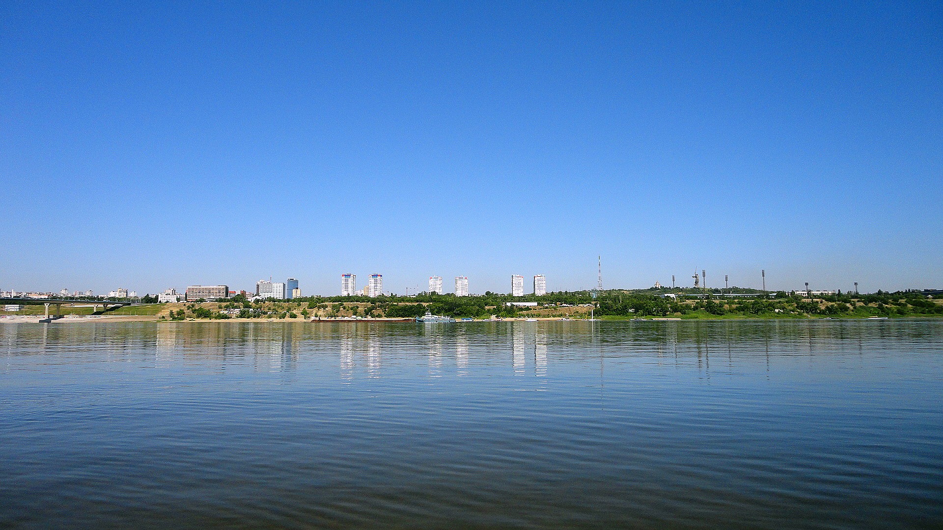 City skyline across a wide river under a clear blue sky HD Wallpaper