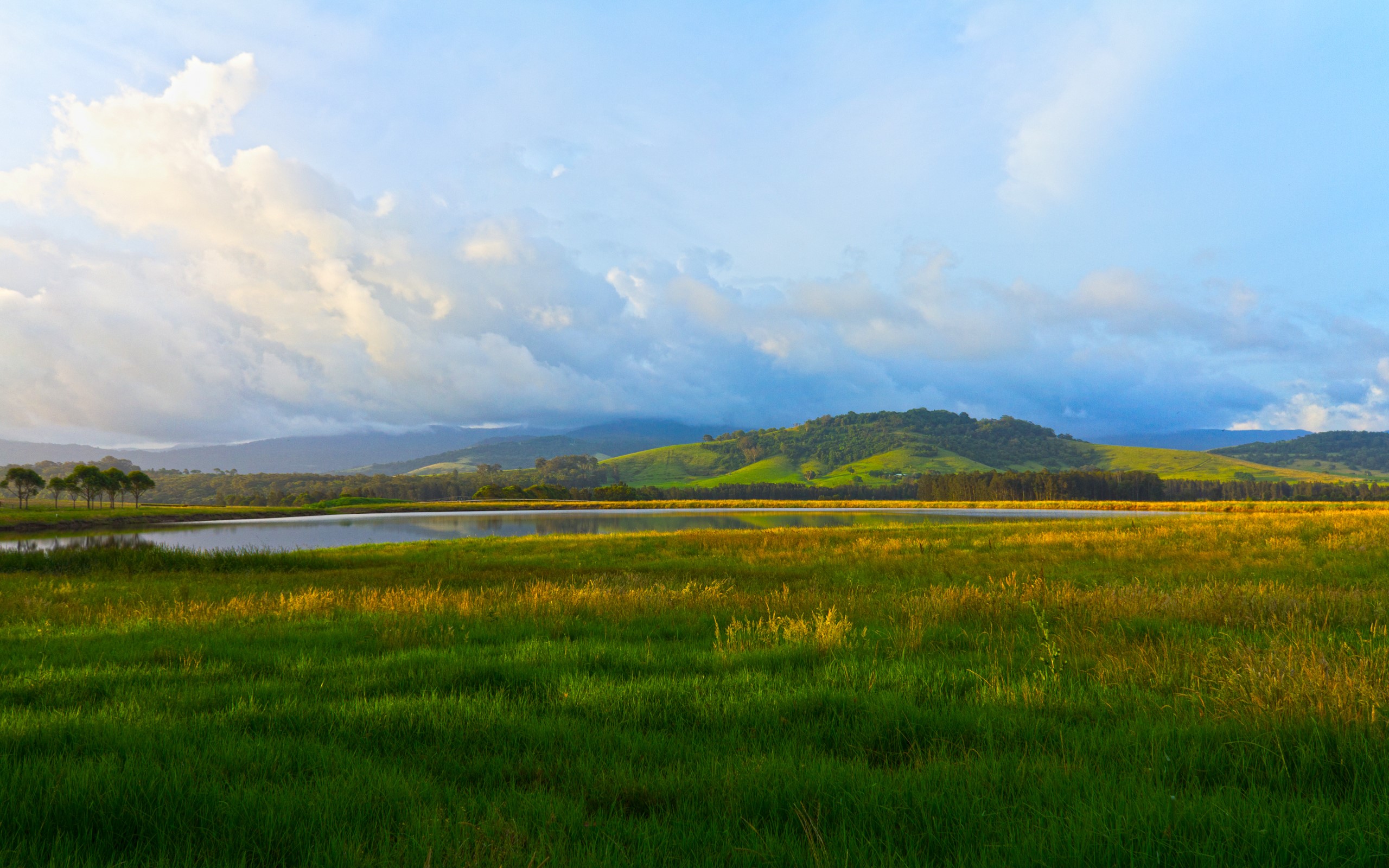 Grassy field with rolling hills and a lake HD Wallpaper