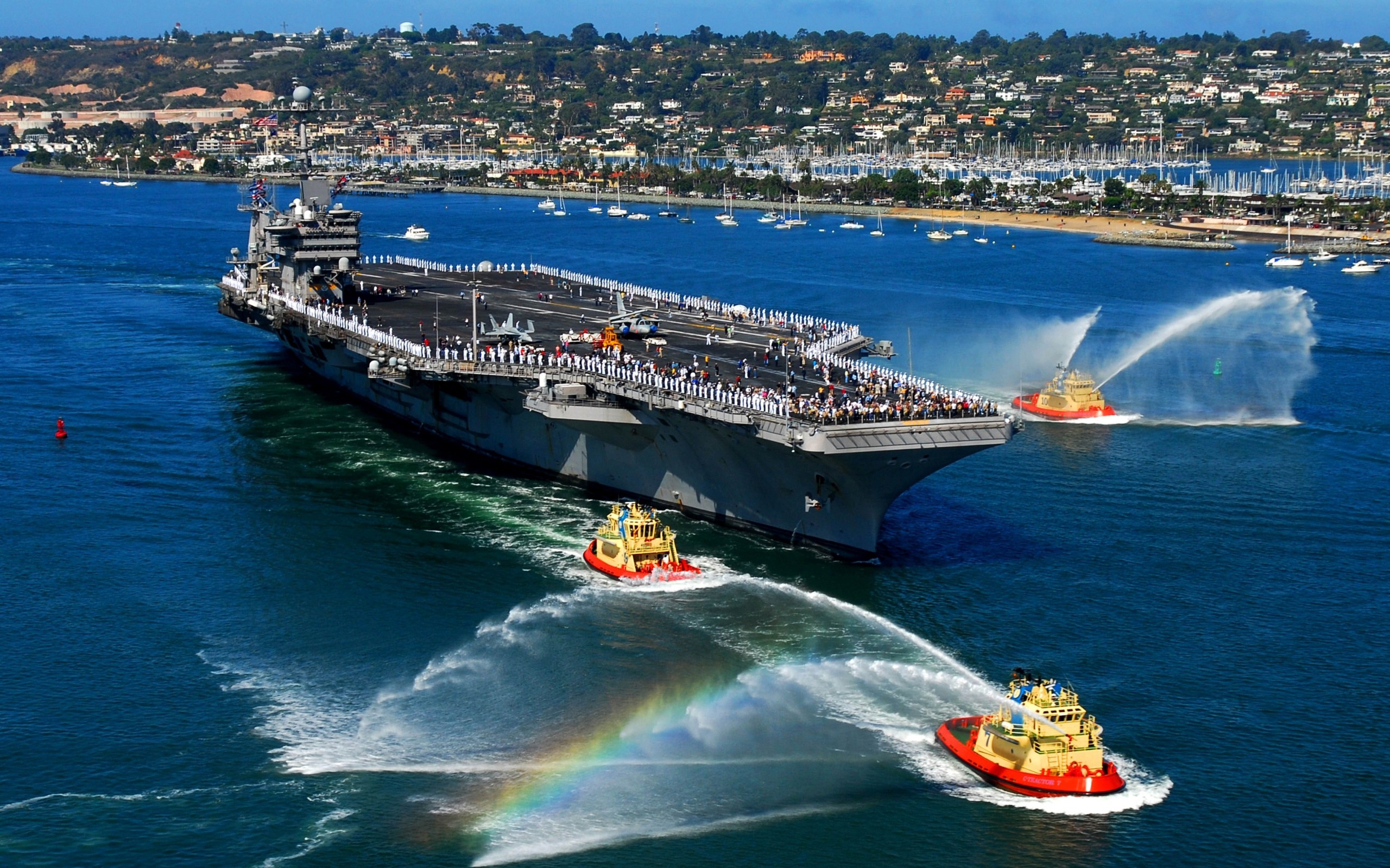 Aircraft carrier entering harbor with tugboats spraying water HD Wallpaper