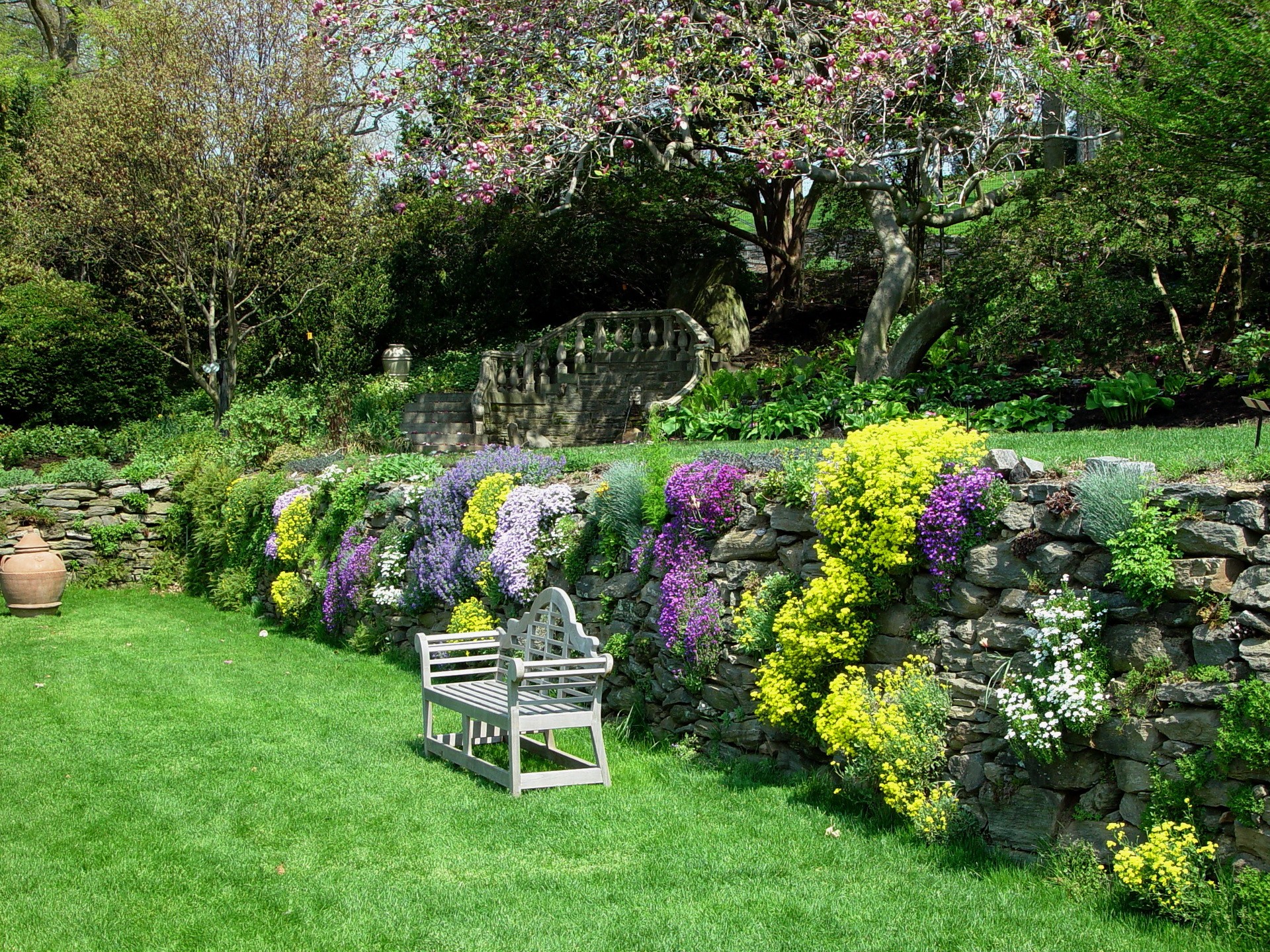 Stone retaining wall with colorful flowers and a bench HD Wallpaper
