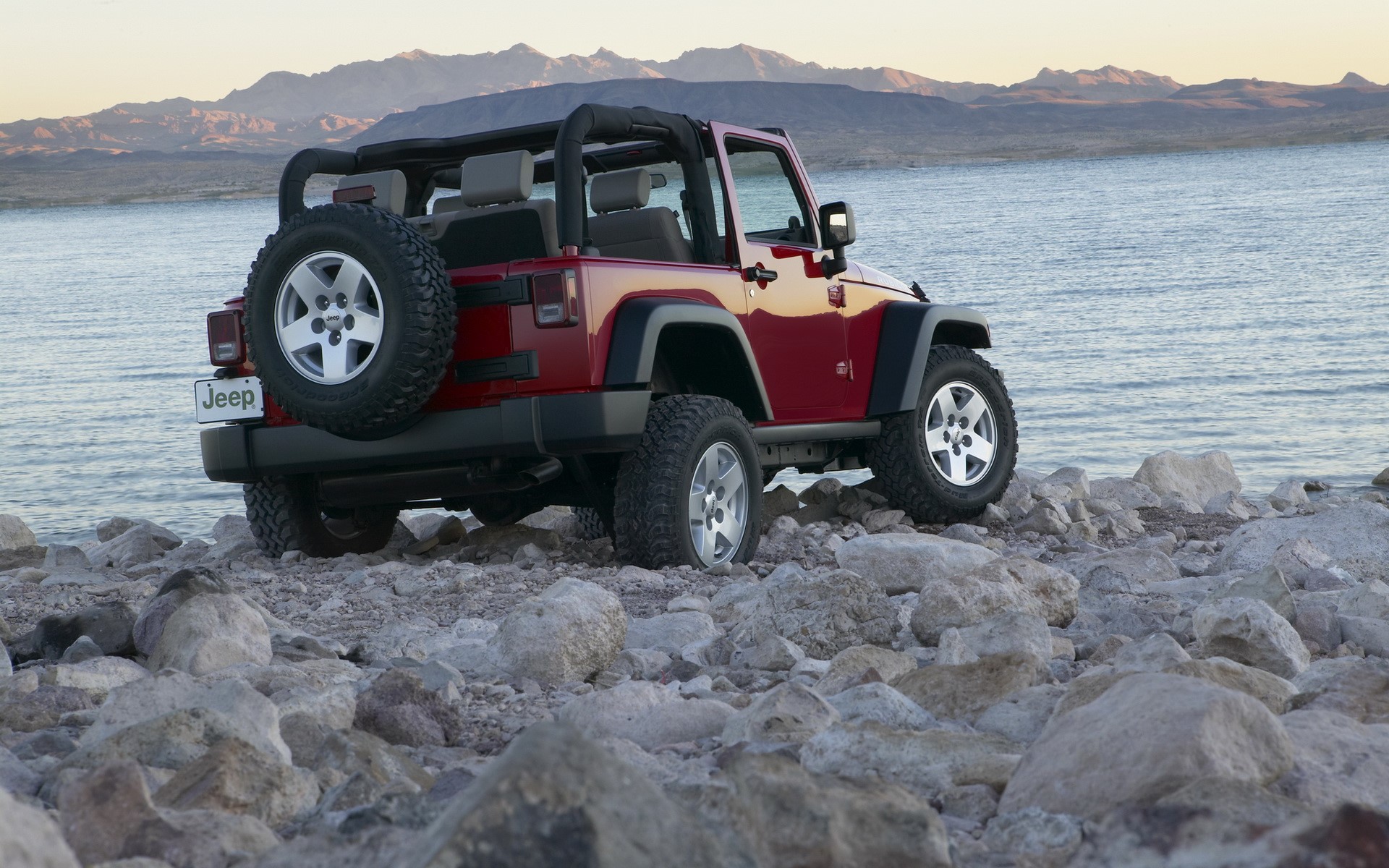 Red Jeep Wrangler parked on rocky shore by lake HD Wallpaper
