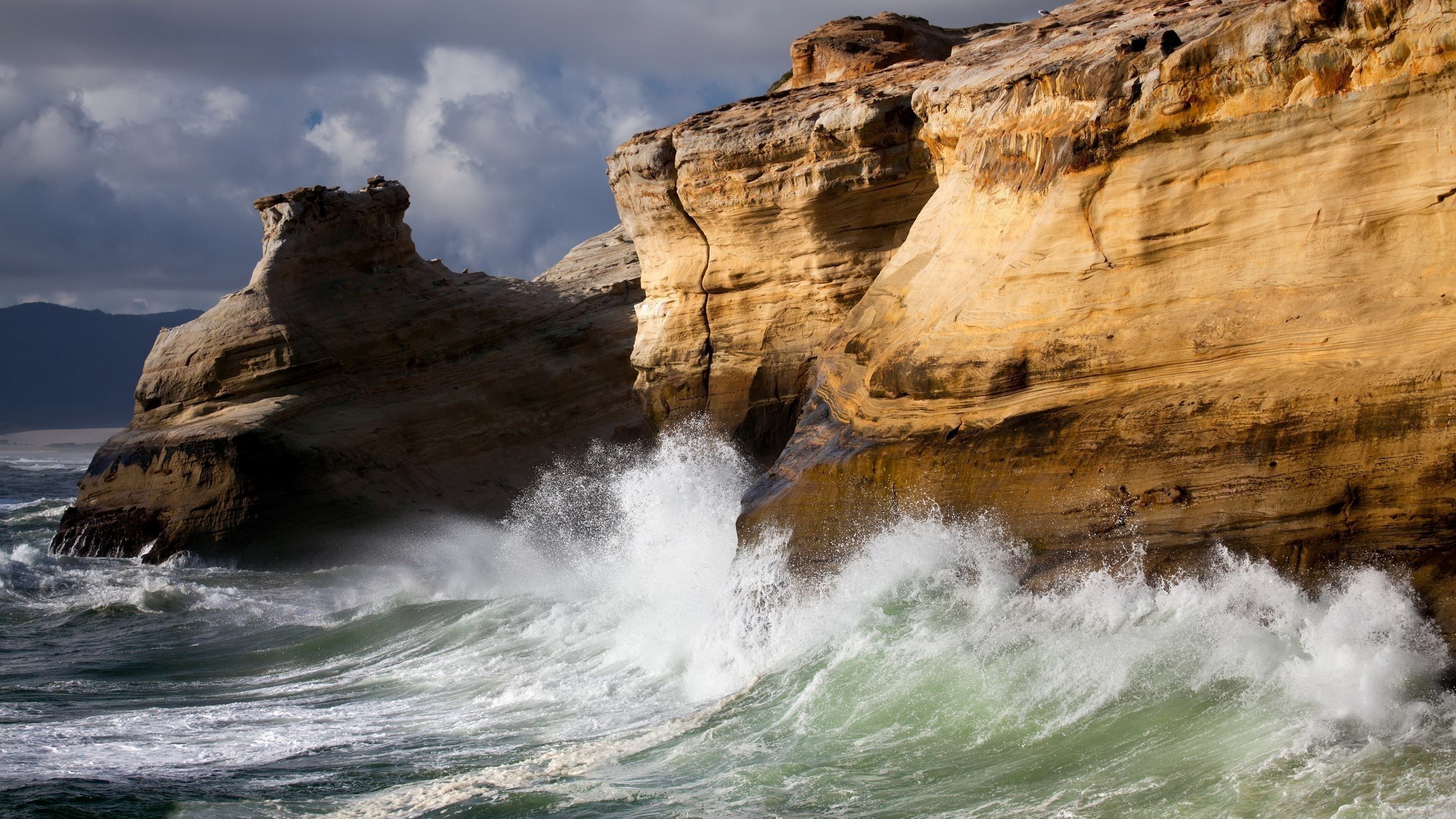 Waves crash against rocky cliffs under a cloudy sky HD Wallpaper