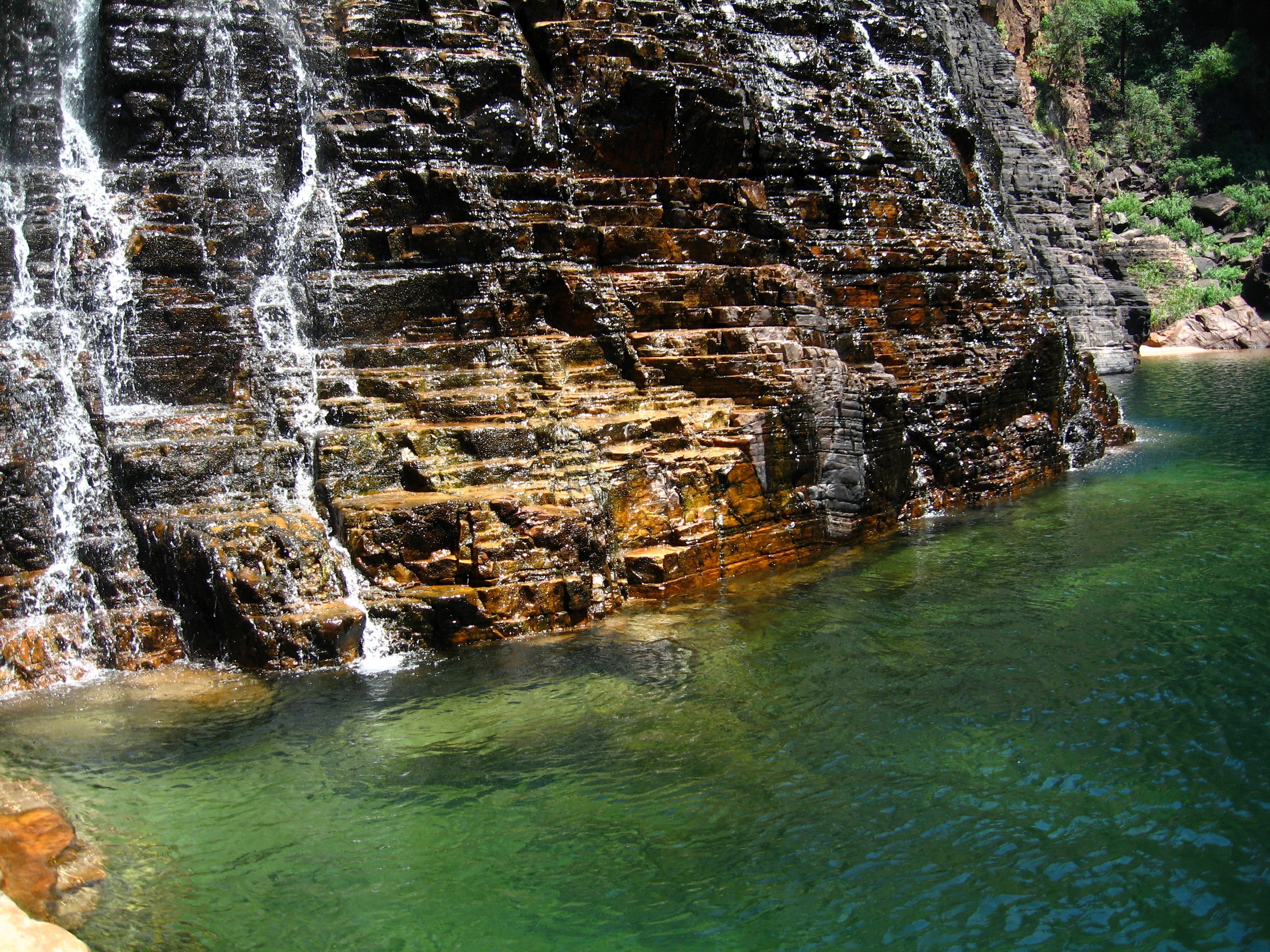Waterfall cascading over layered rock into a clear green pool HD Wallpaper
