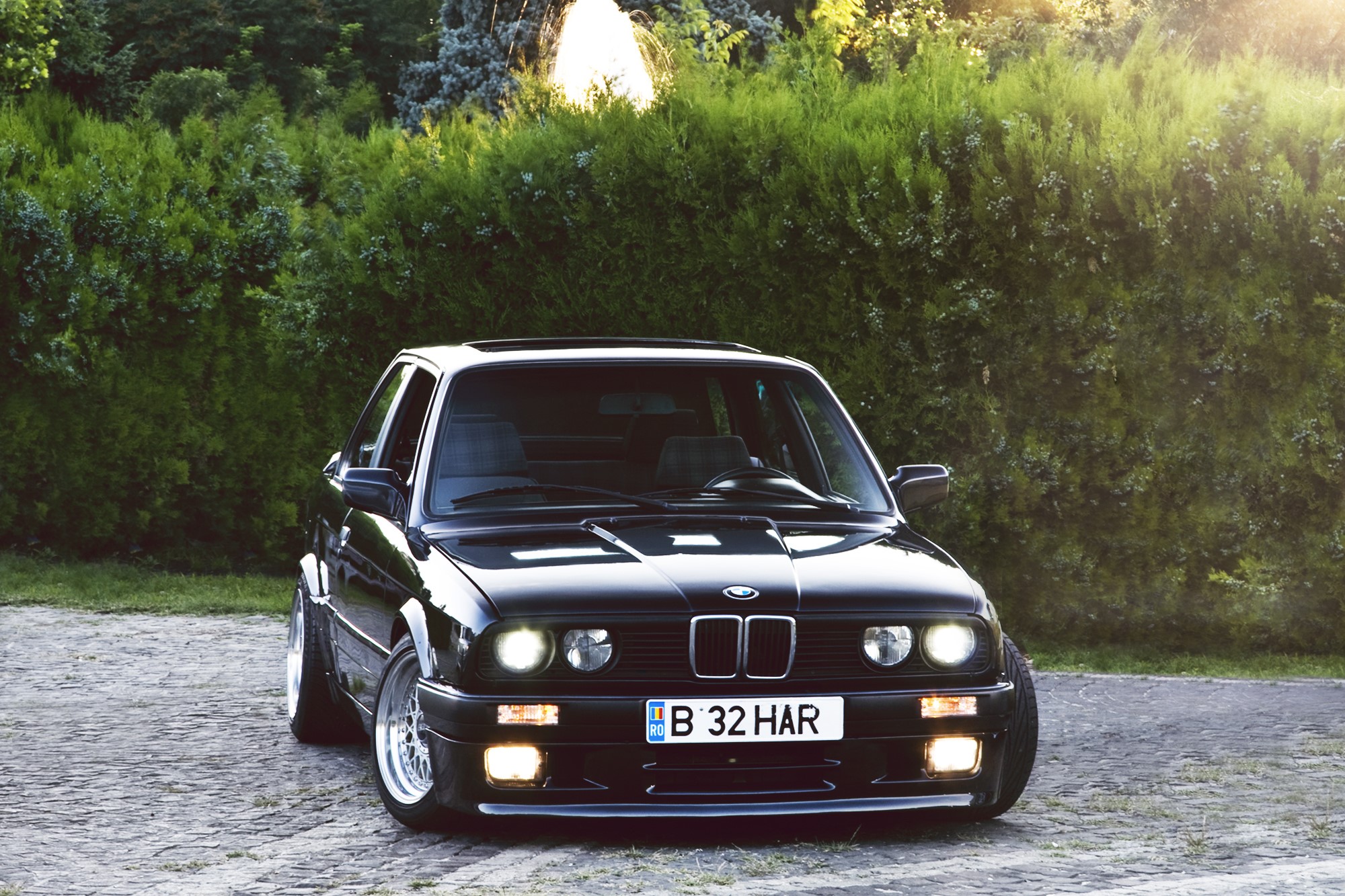 Black BMW E30 parked on cobblestone with trees in background HD Wallpaper