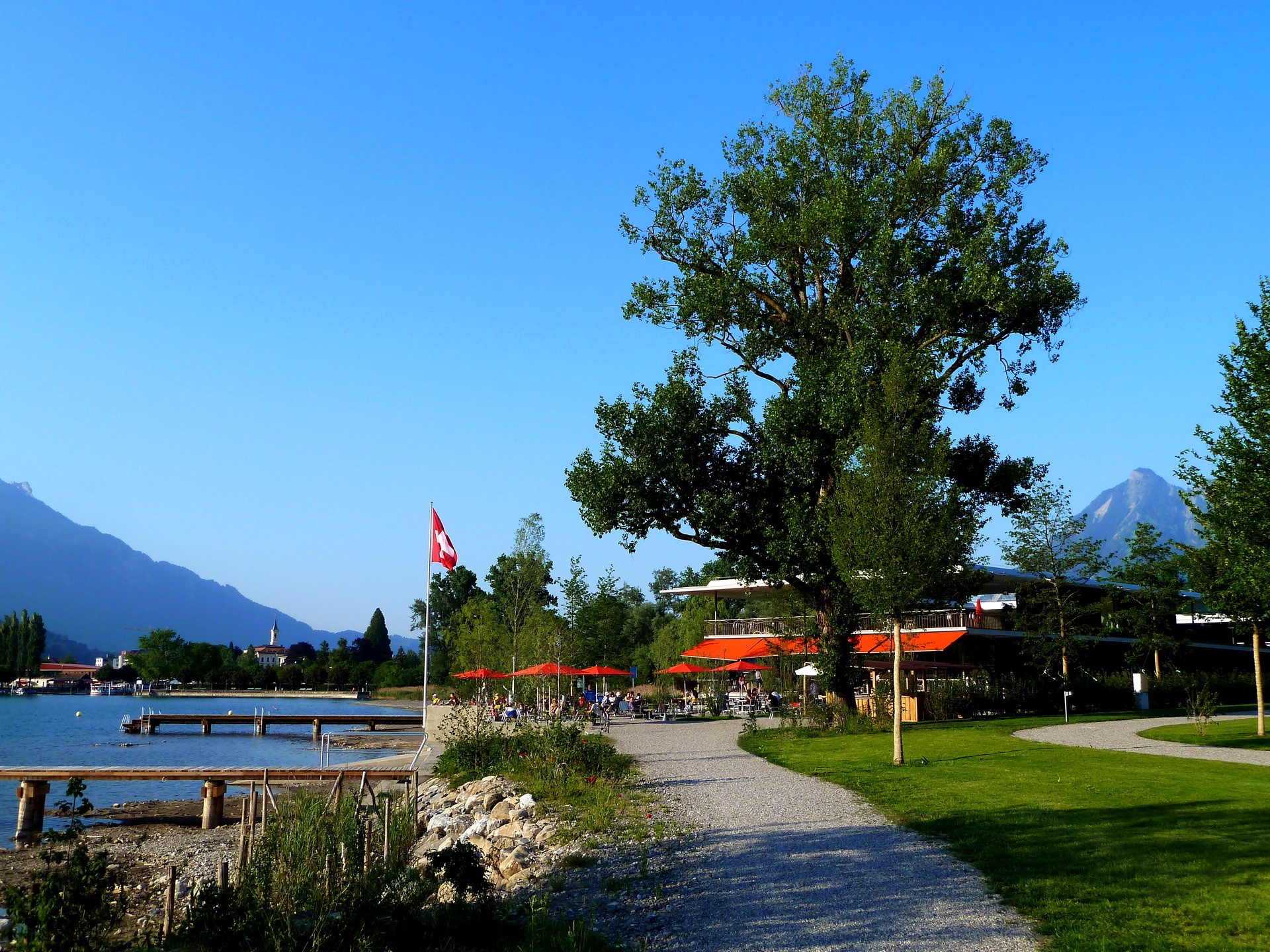 Swiss flag by a lake with mountains and a building HD Wallpaper
