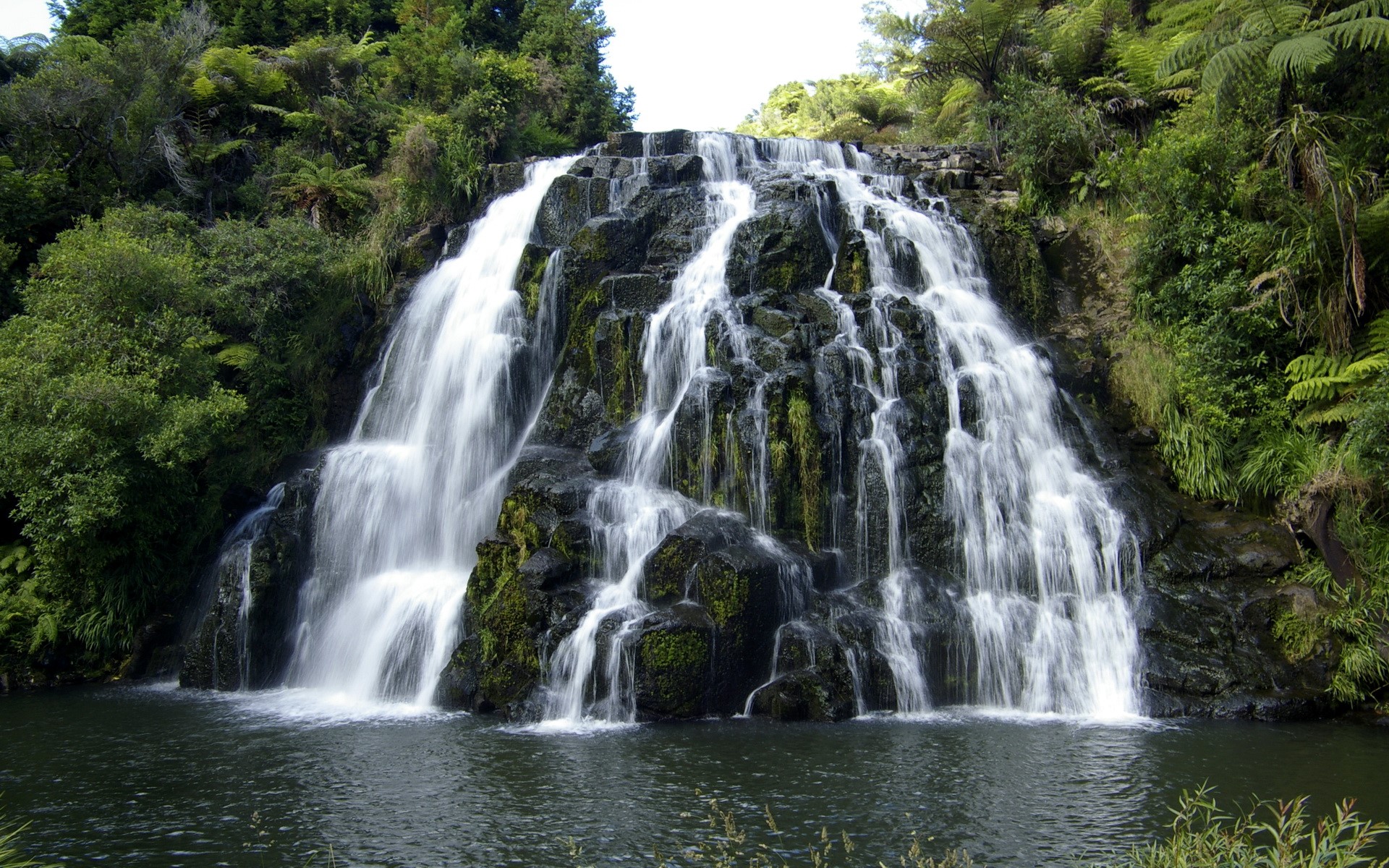 A wide waterfall cascades over moss-covered rocks into a pool below. HD Wallpaper