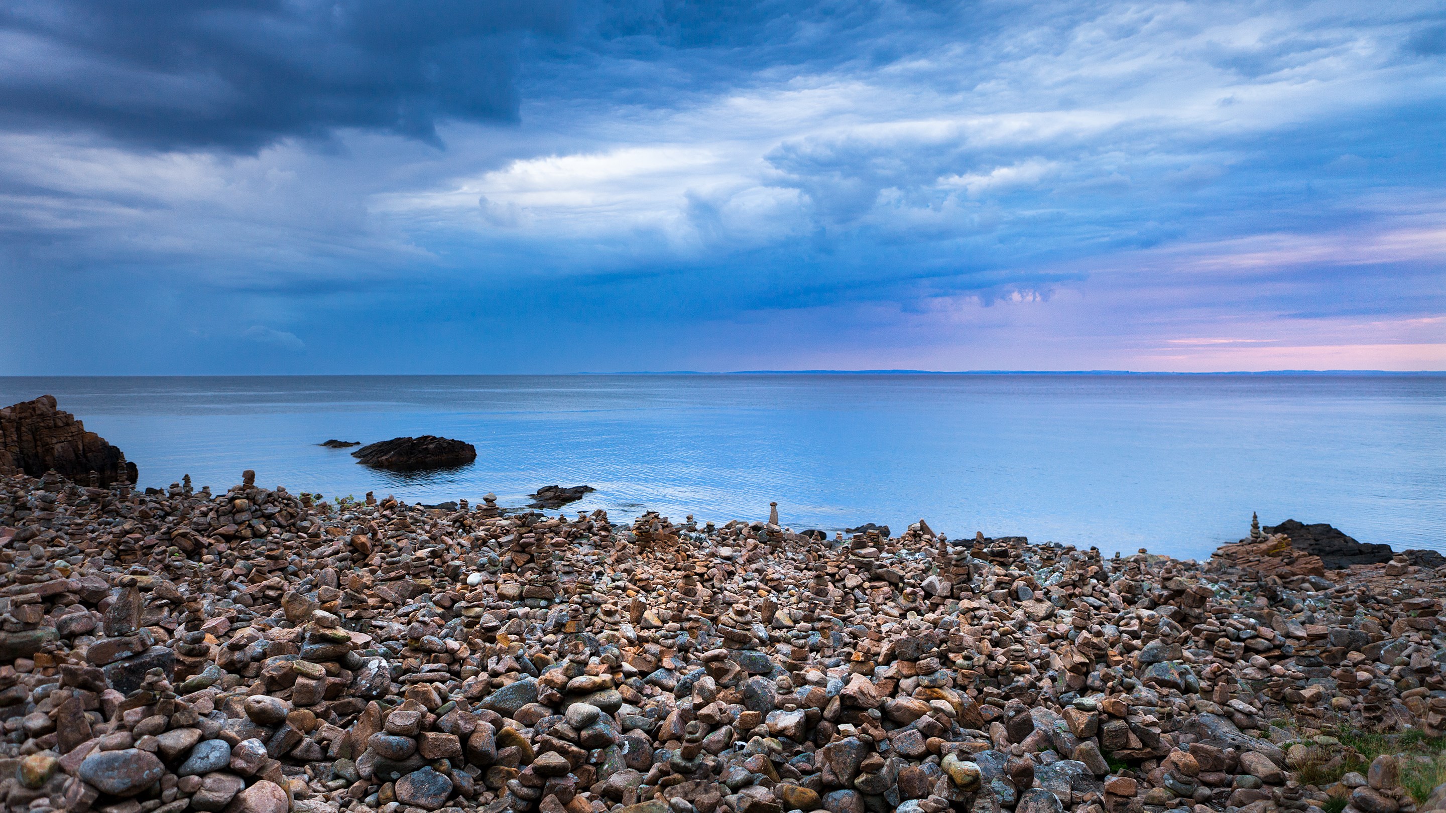 Rocky shore with stacked stones and ocean under cloudy sky HD Wallpaper