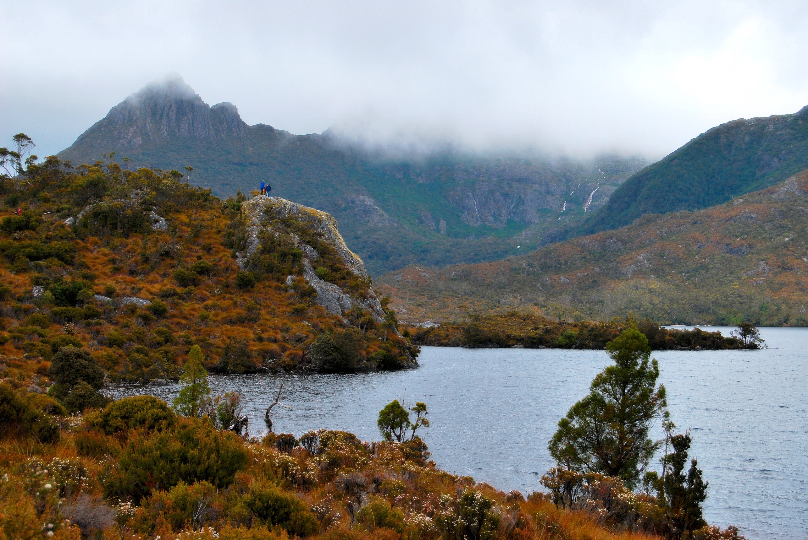 Two hikers on a rocky outcrop overlooking a lake in Tasmania, Australia HD Wallpaper