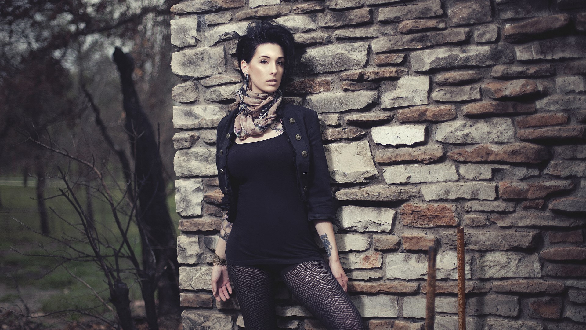 Woman in black dress and patterned pantyhose leaning against stone wall HD Wallpaper