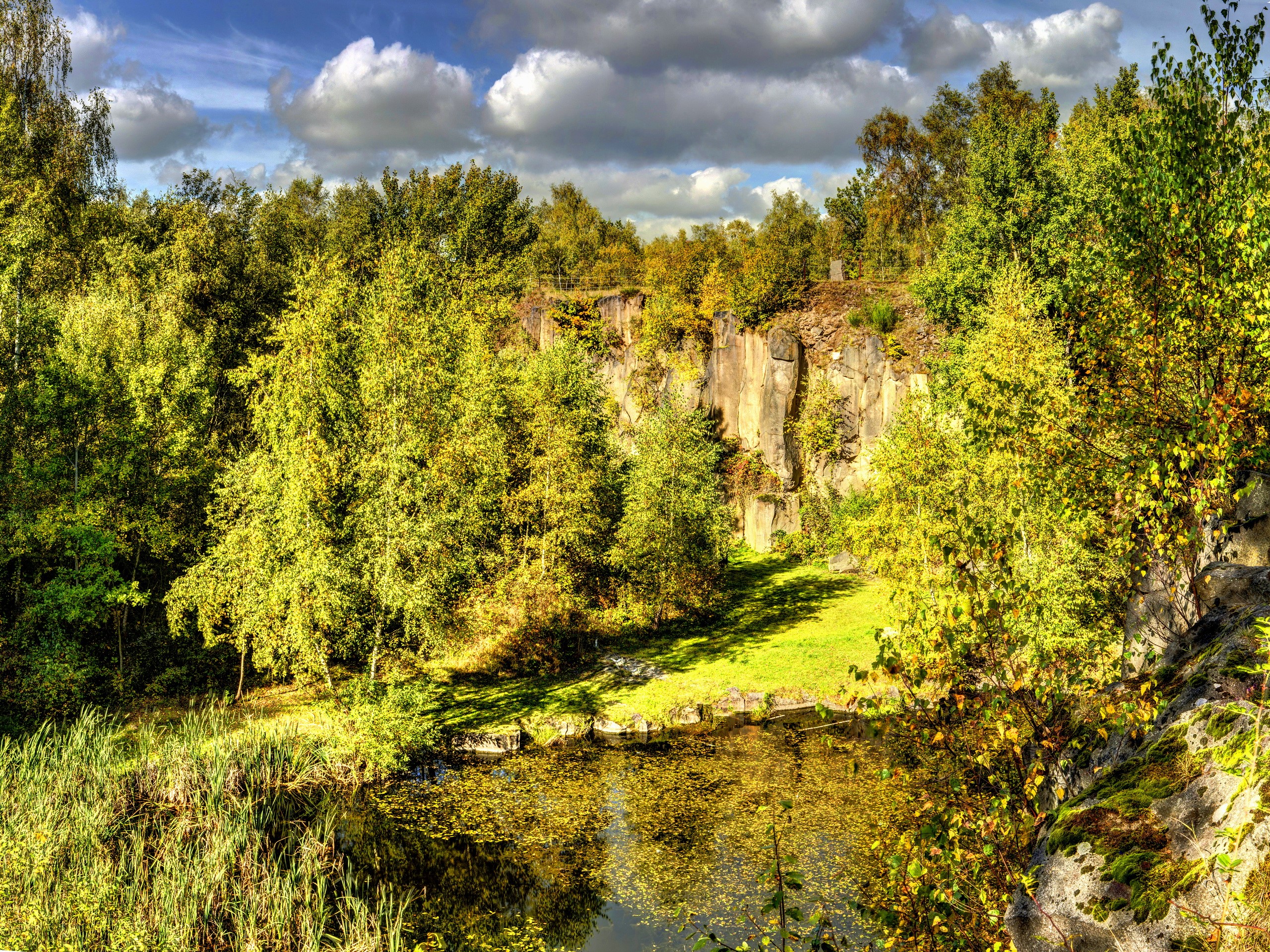 Quarry lake with trees and rocky cliffs under clouds HD Wallpaper