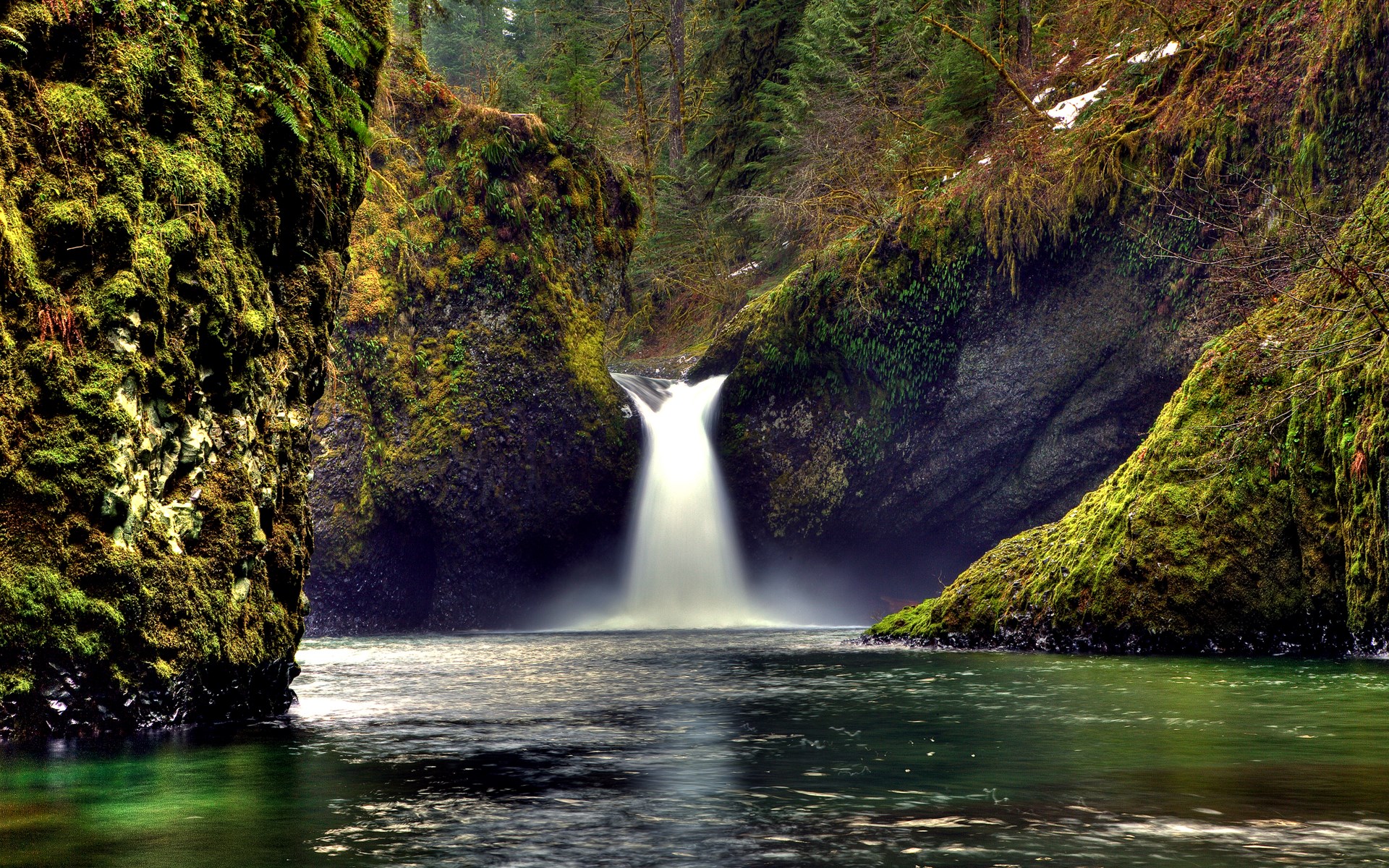 Moss-covered rocks surround a waterfall flowing into a river HD Wallpaper