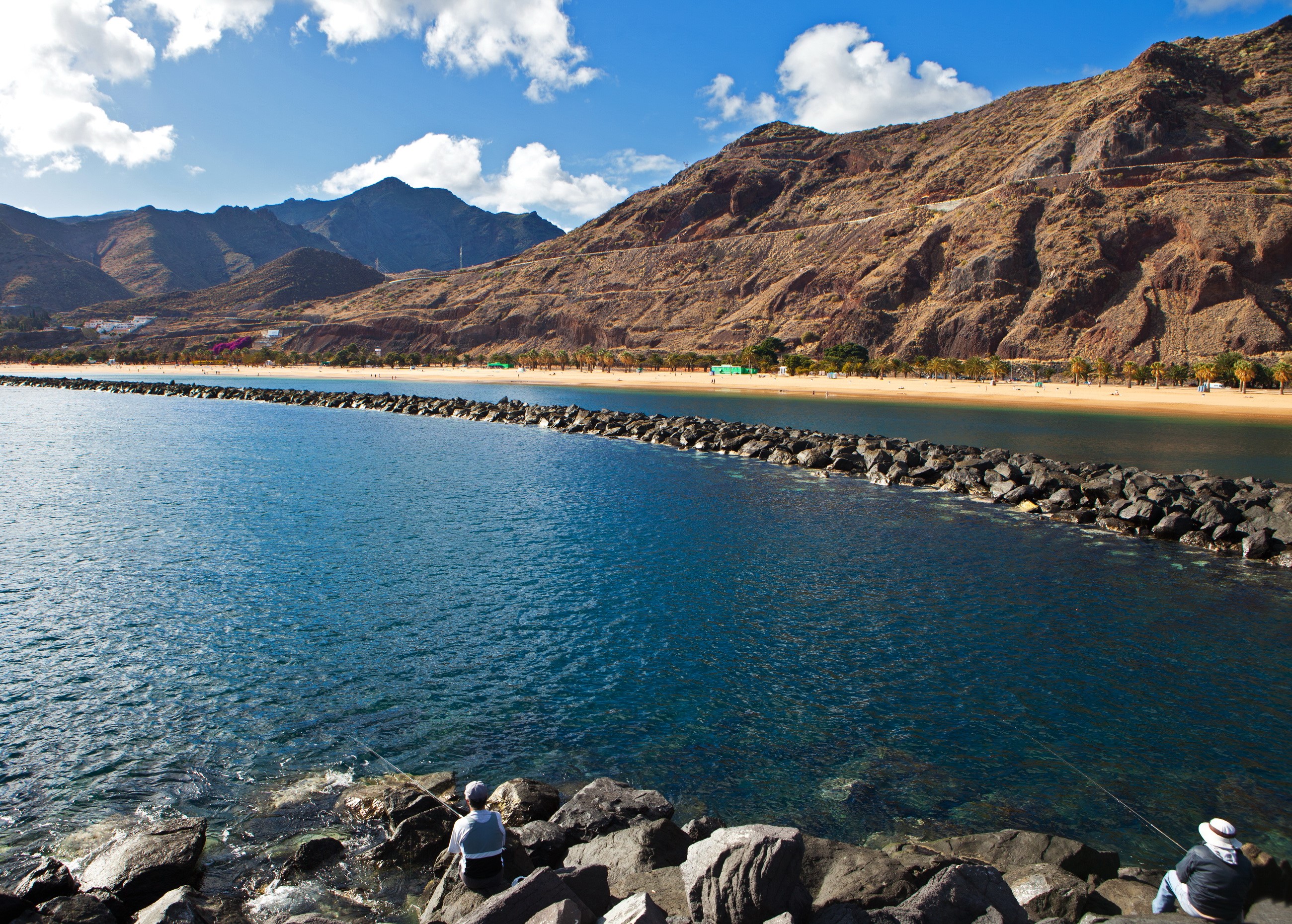 Two people fishing on rocks with a beach and mountains in the background HD Wallpaper