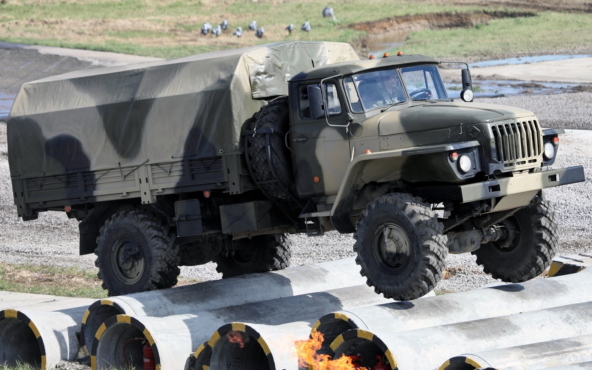 Military truck driving over concrete pipes with fire HD Wallpaper
