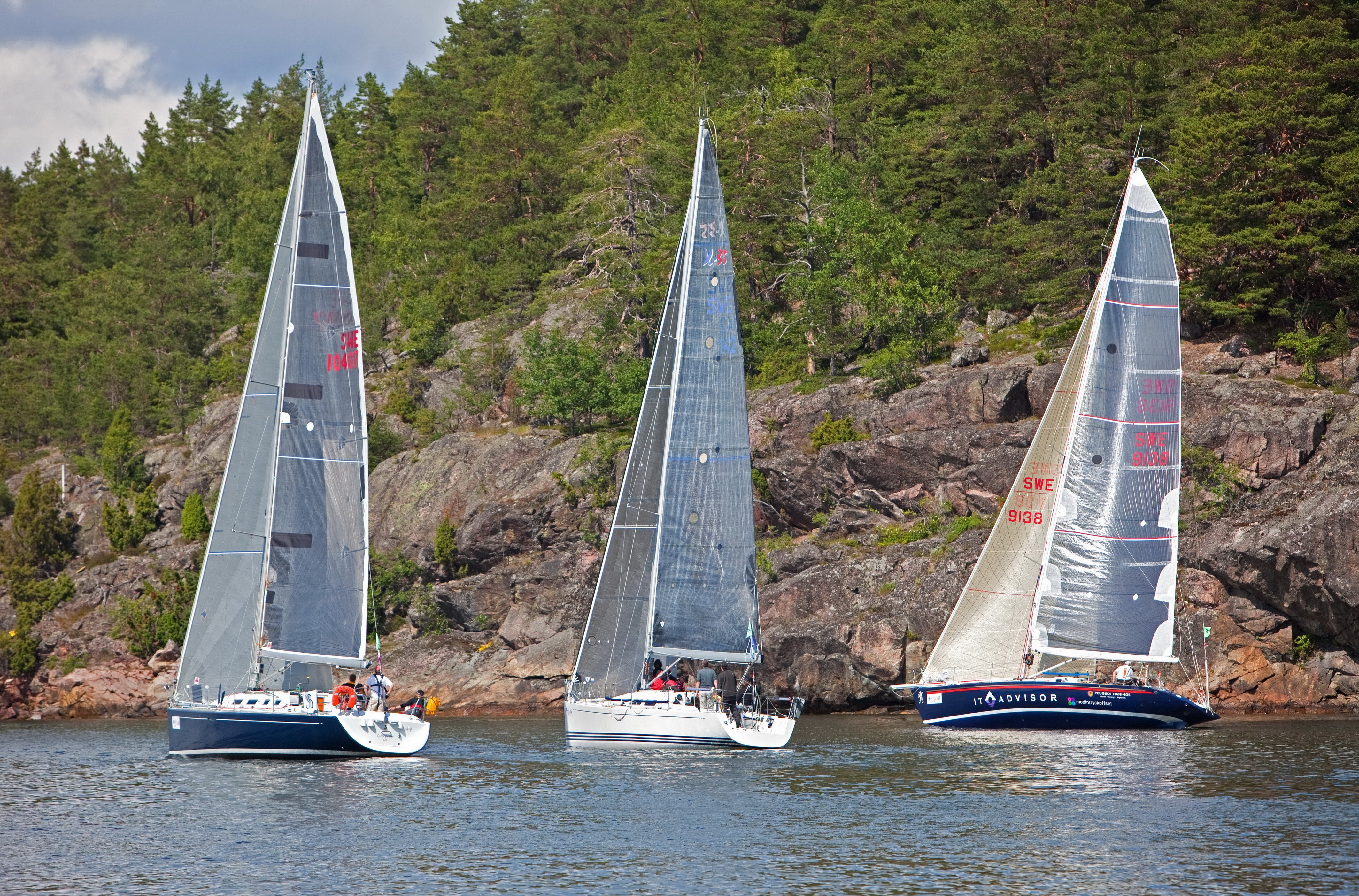 Three sailboats racing near a rocky, tree-covered shore HD Wallpaper