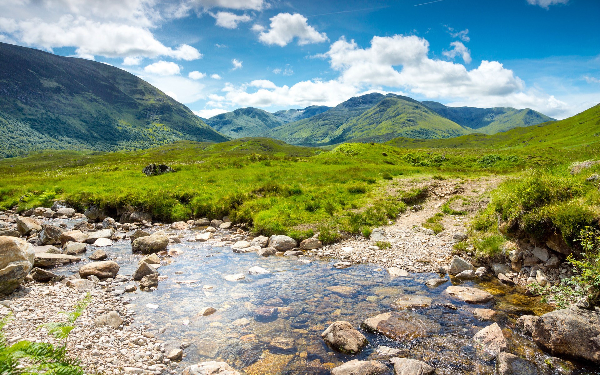 A clear stream flows through a green valley with mountains HD Wallpaper