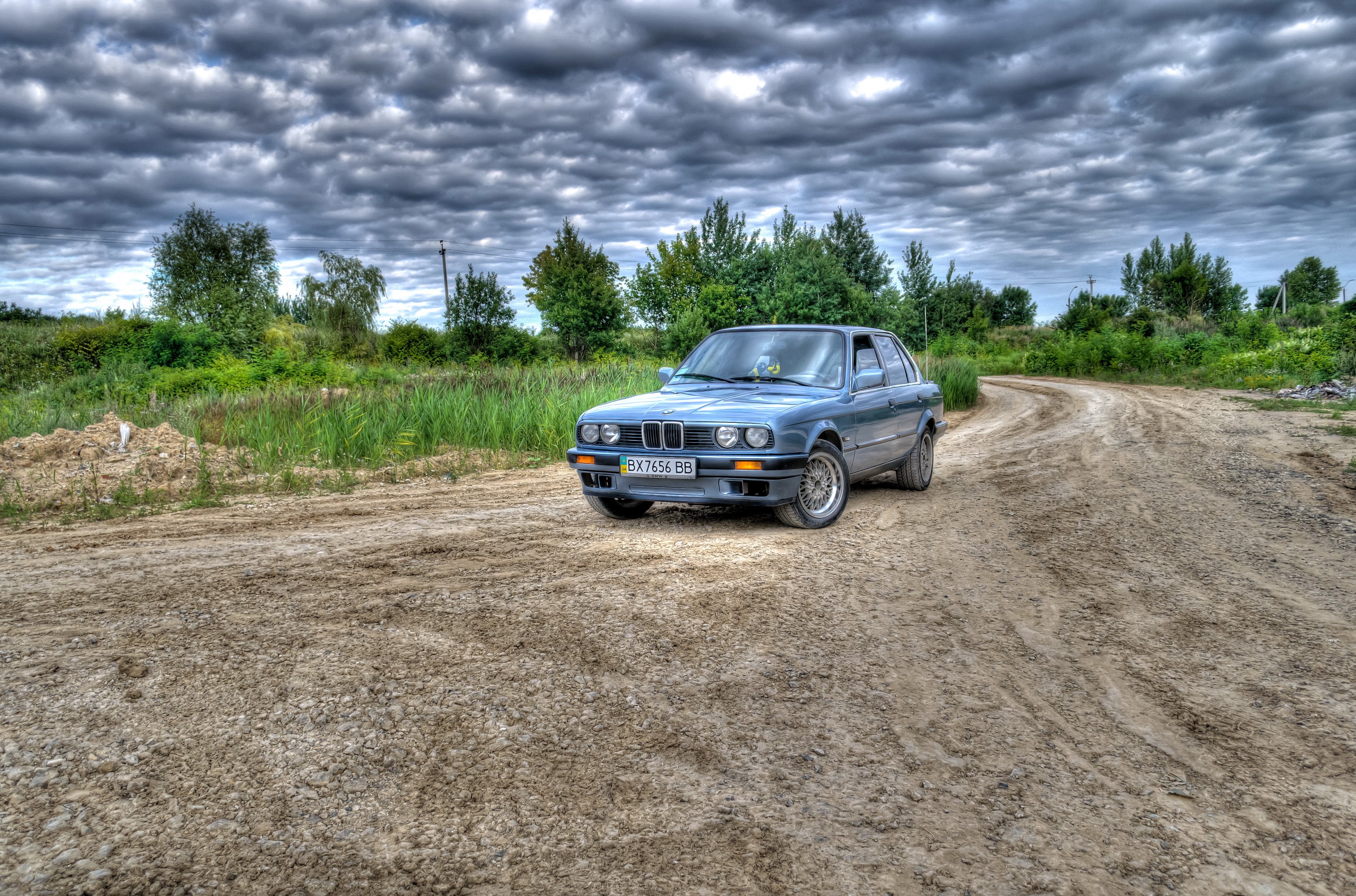 Blue BMW on a dirt road under cloudy sky HD Wallpaper