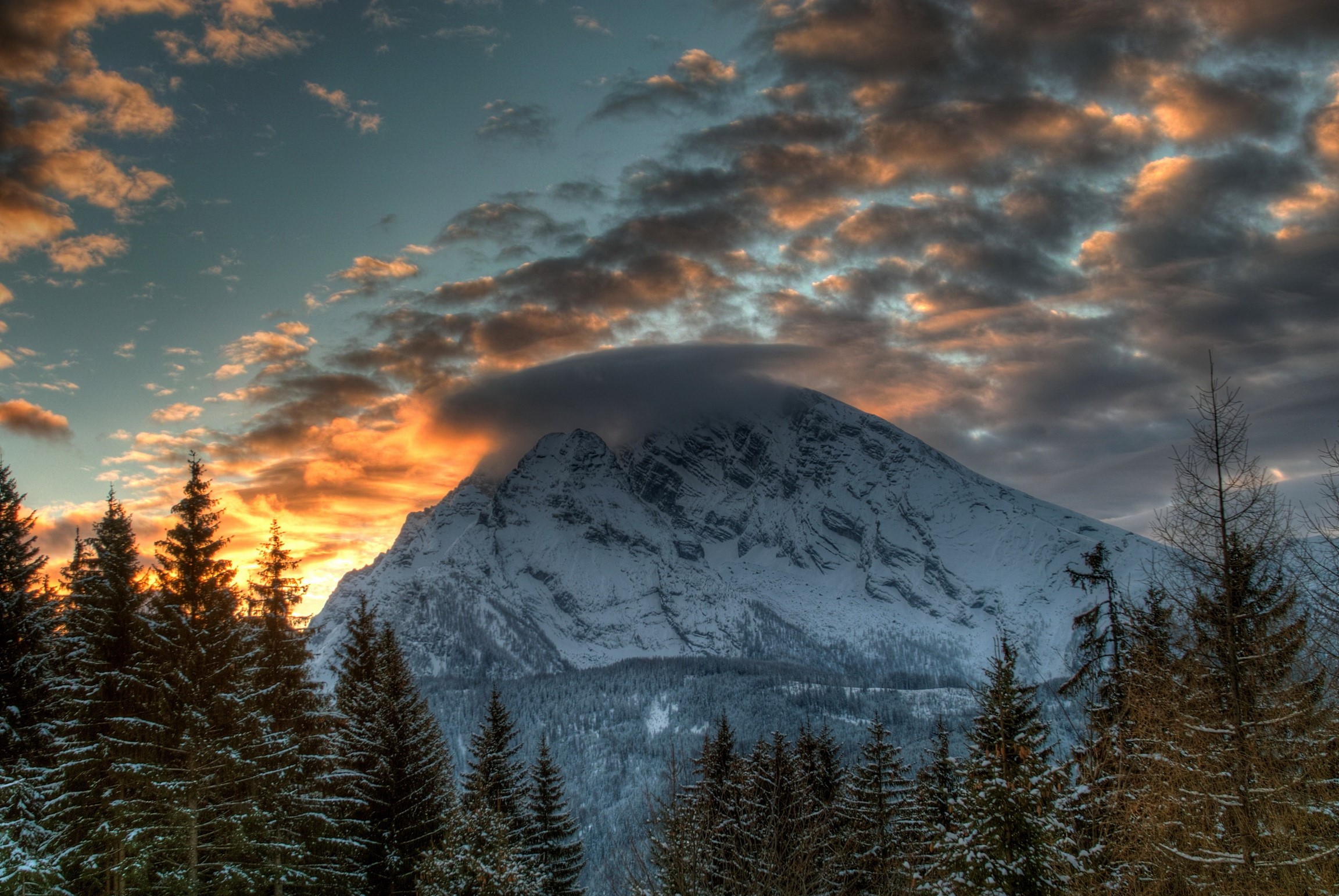 Snowy mountain peak with dramatic sunset clouds and pine forest HD Wallpaper