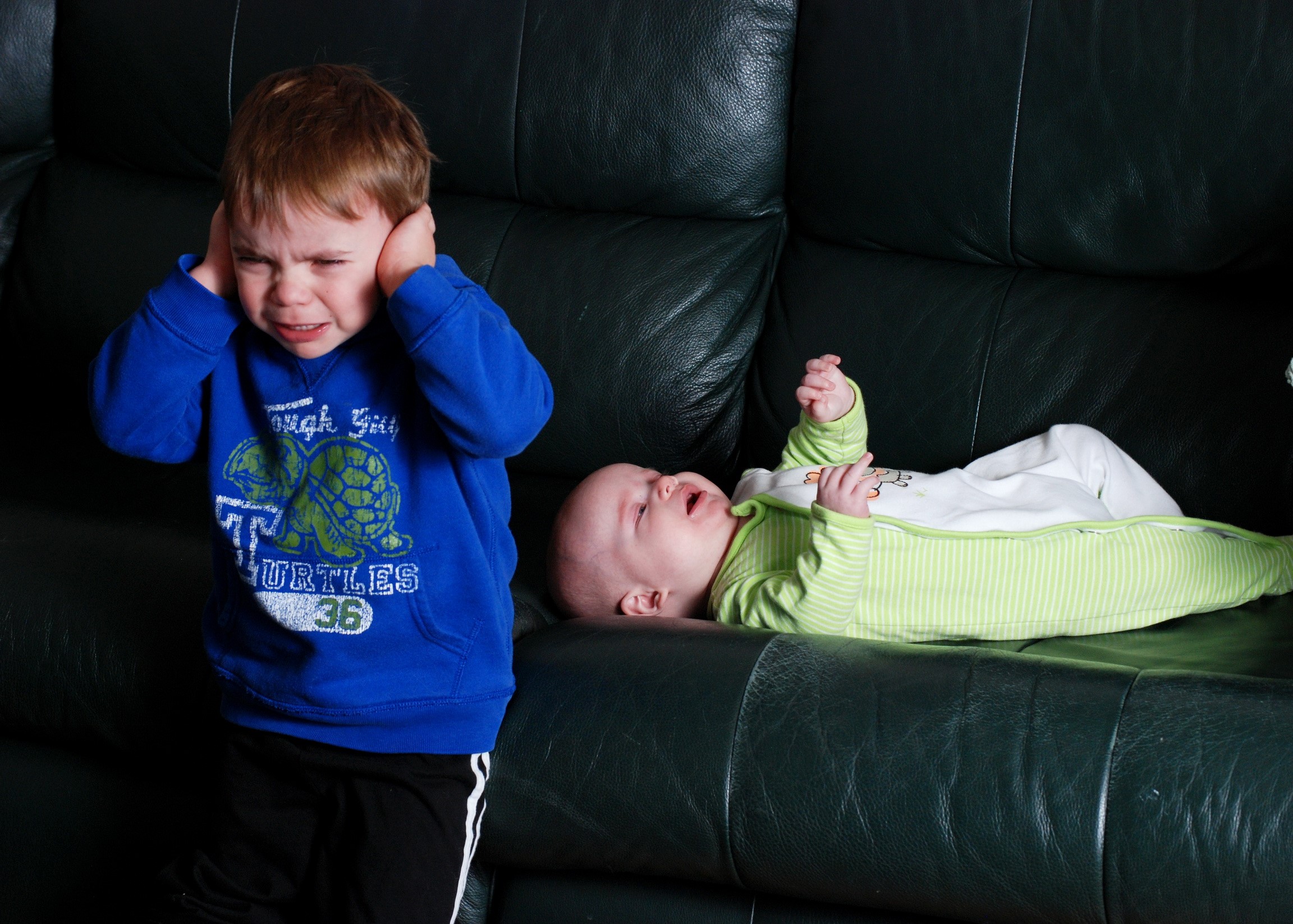 Boy covering ears next to crying baby on couch HD Wallpaper