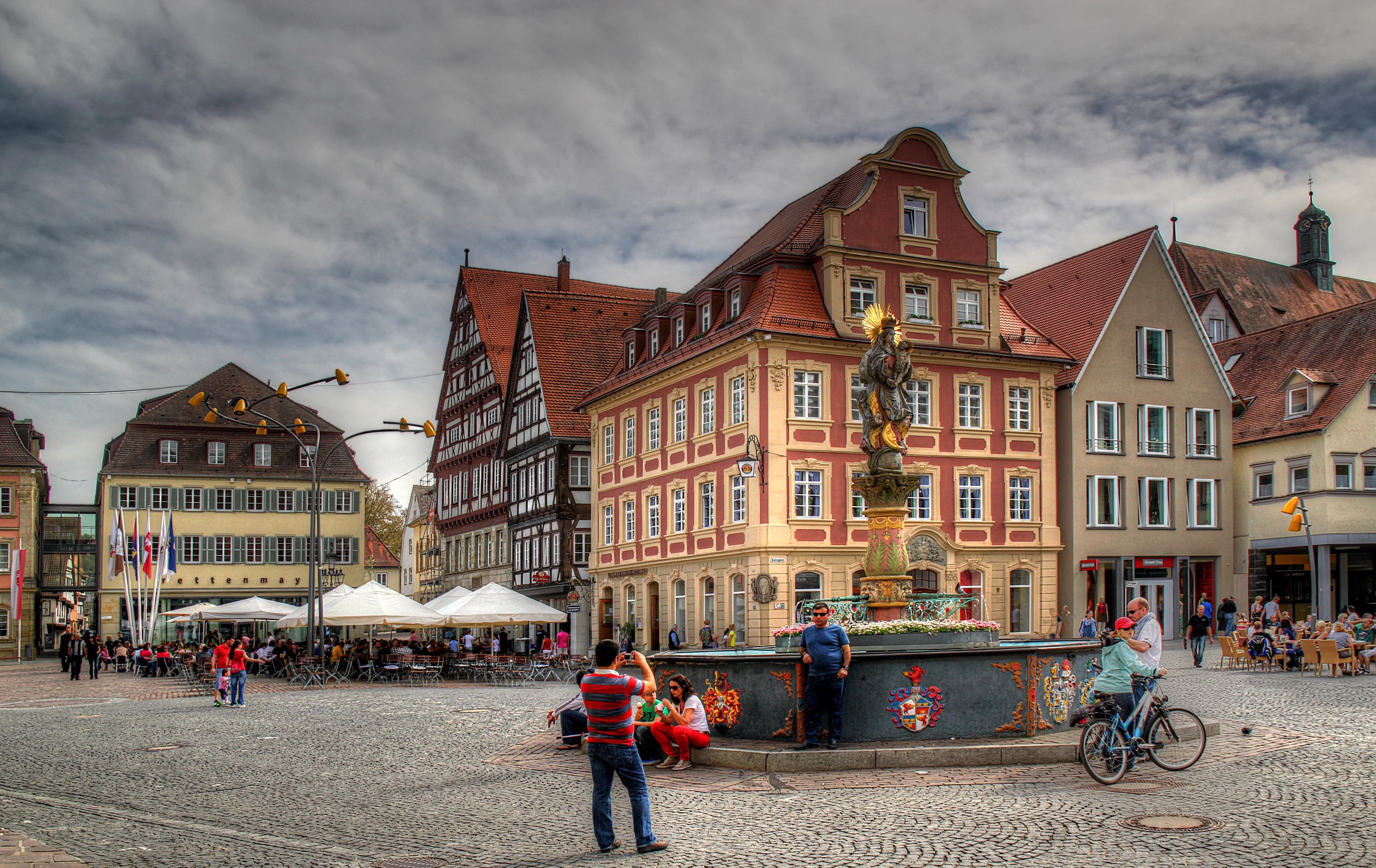 People in a European town square with a fountain HD Wallpaper