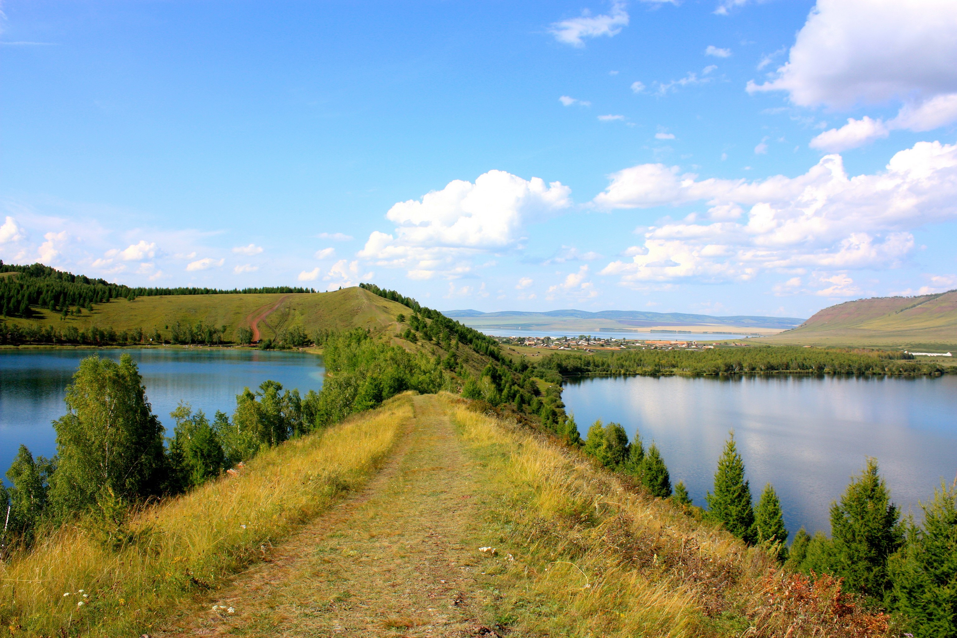 A scenic view of a lake with rolling hills and a path HD Wallpaper