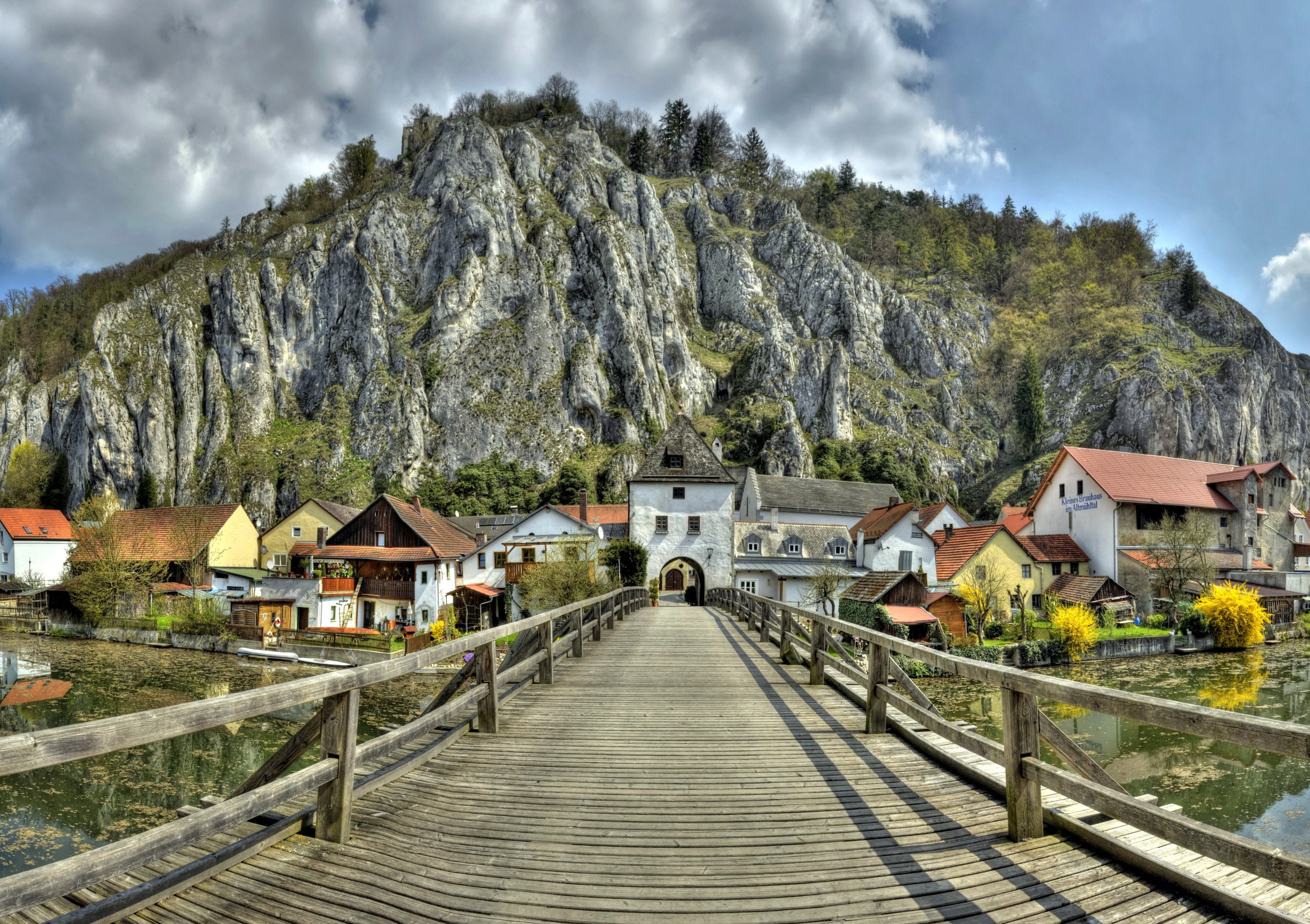 Wooden bridge leading to Essing, Germany with a rocky mountain backdrop HD Wallpaper