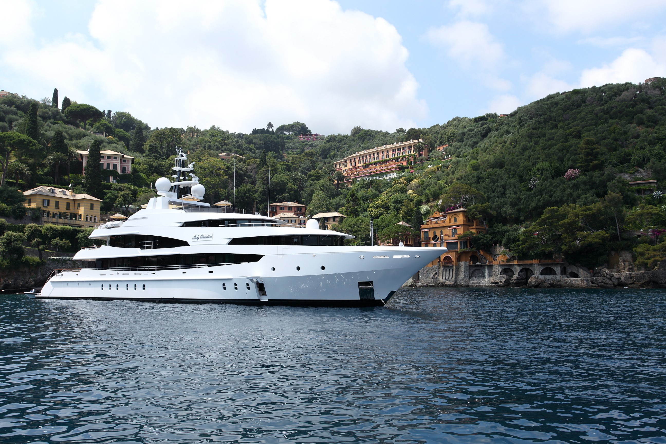 Luxury yacht anchored near a lush green hillside with villas HD Wallpaper