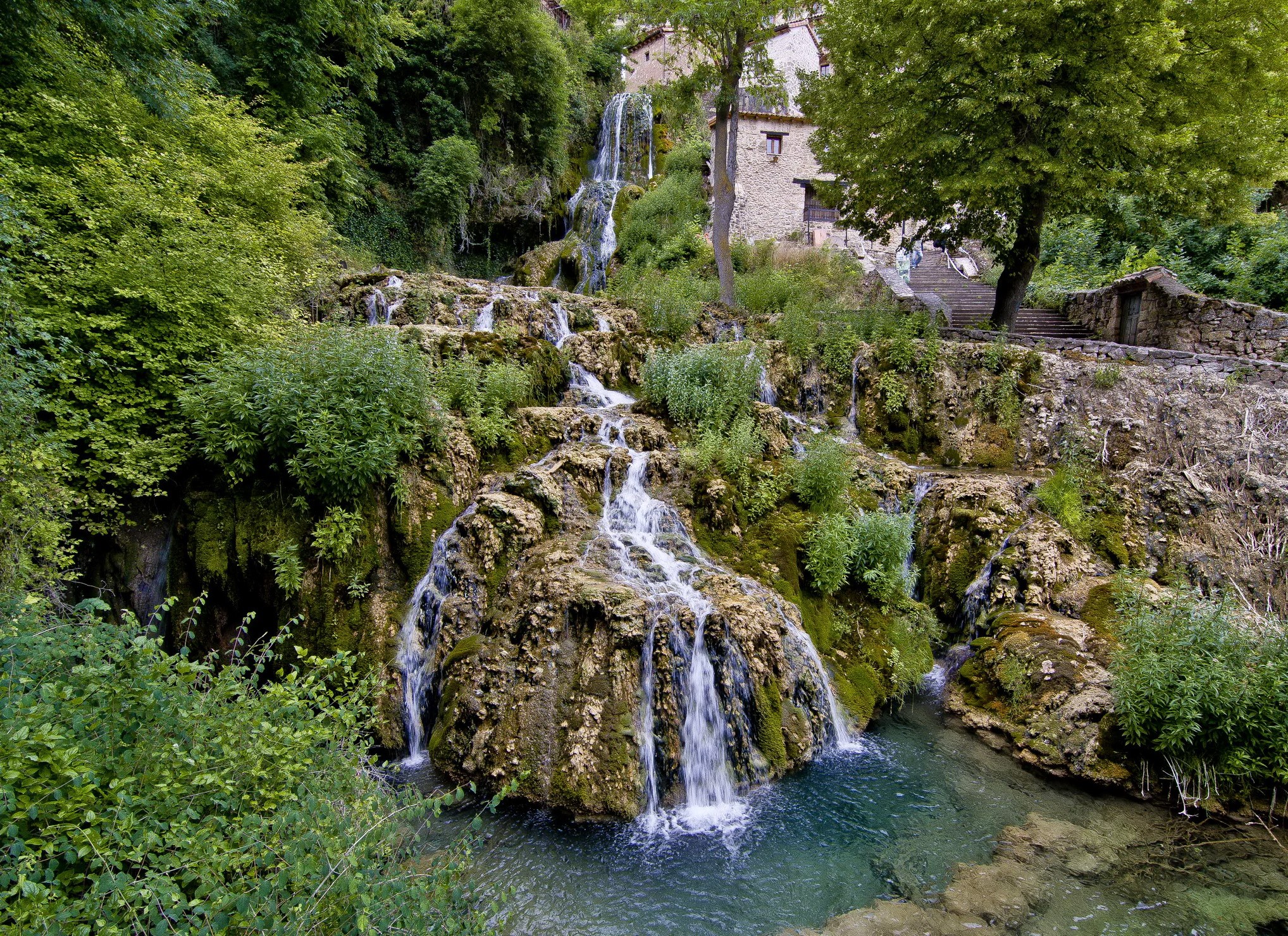 Waterfalls cascading down mossy rocks near a stone building in Burgos, Spain HD Wallpaper