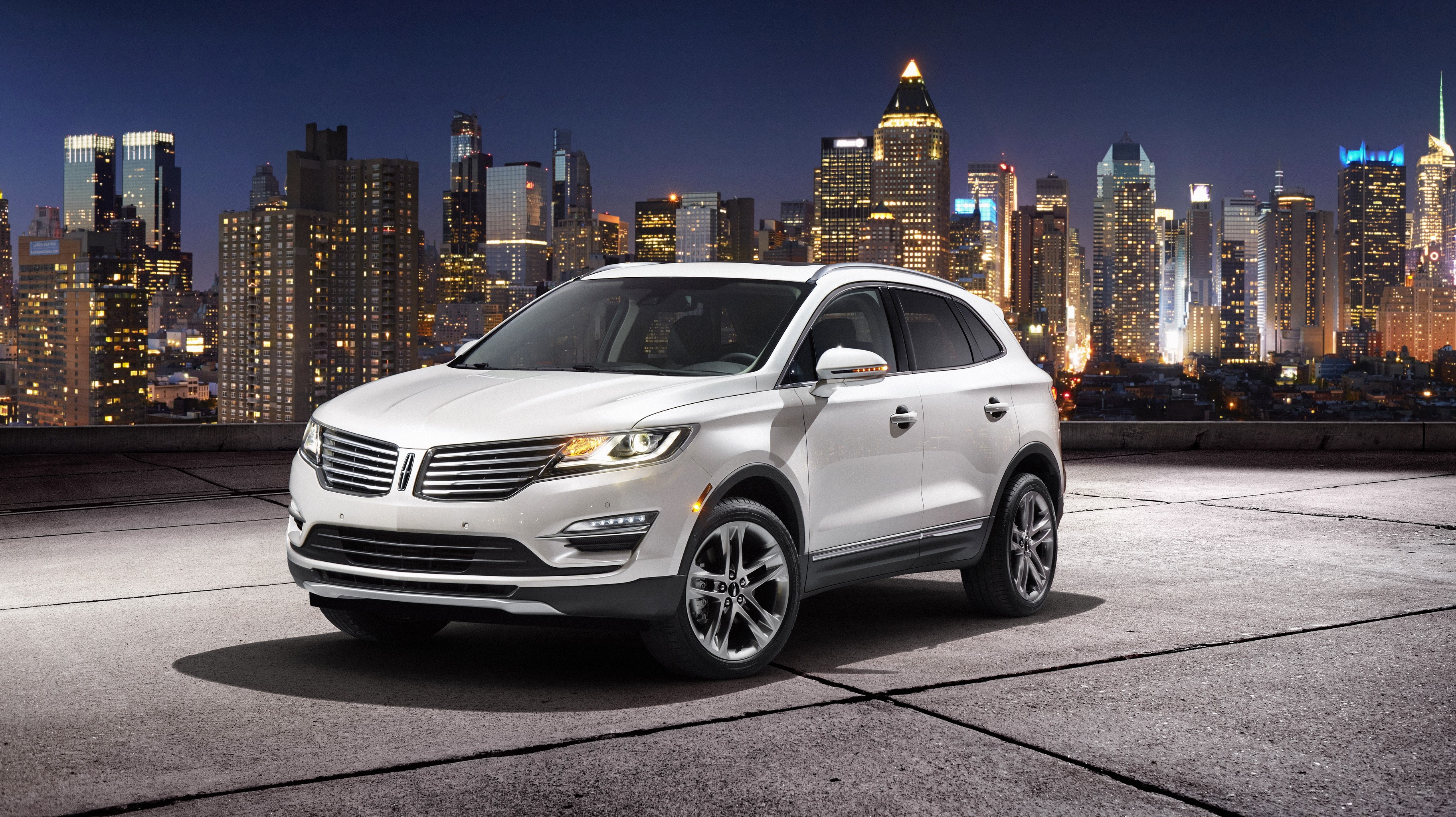 White Lincoln MKC parked in front of a city skyline at night HD Wallpaper