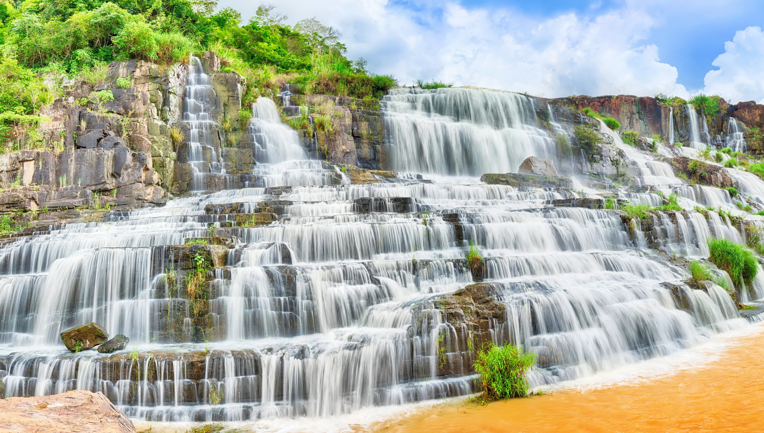 Multi-tiered waterfall cascading over rocks with lush greenery HD Wallpaper