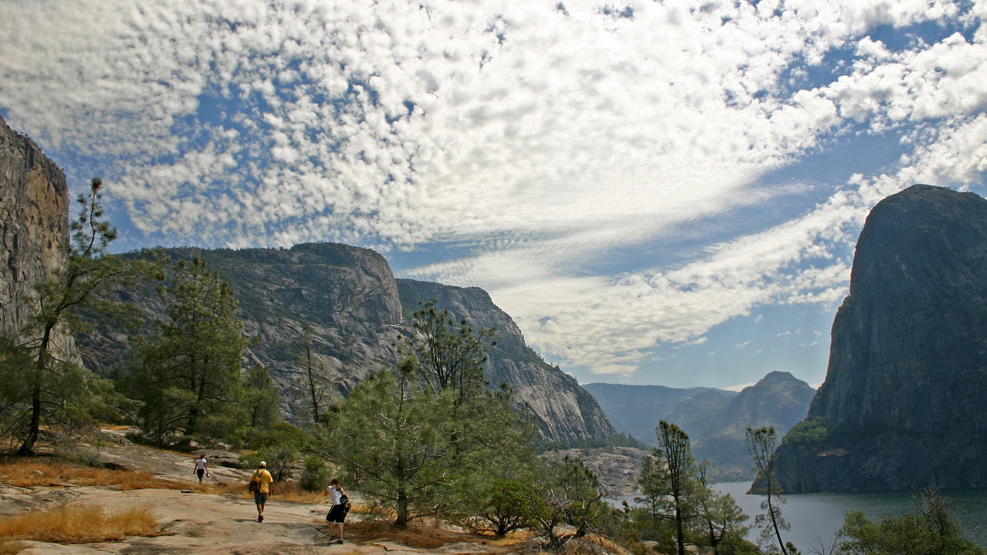 Hikers on a rocky trail with granite cliffs and a lake HD Wallpaper