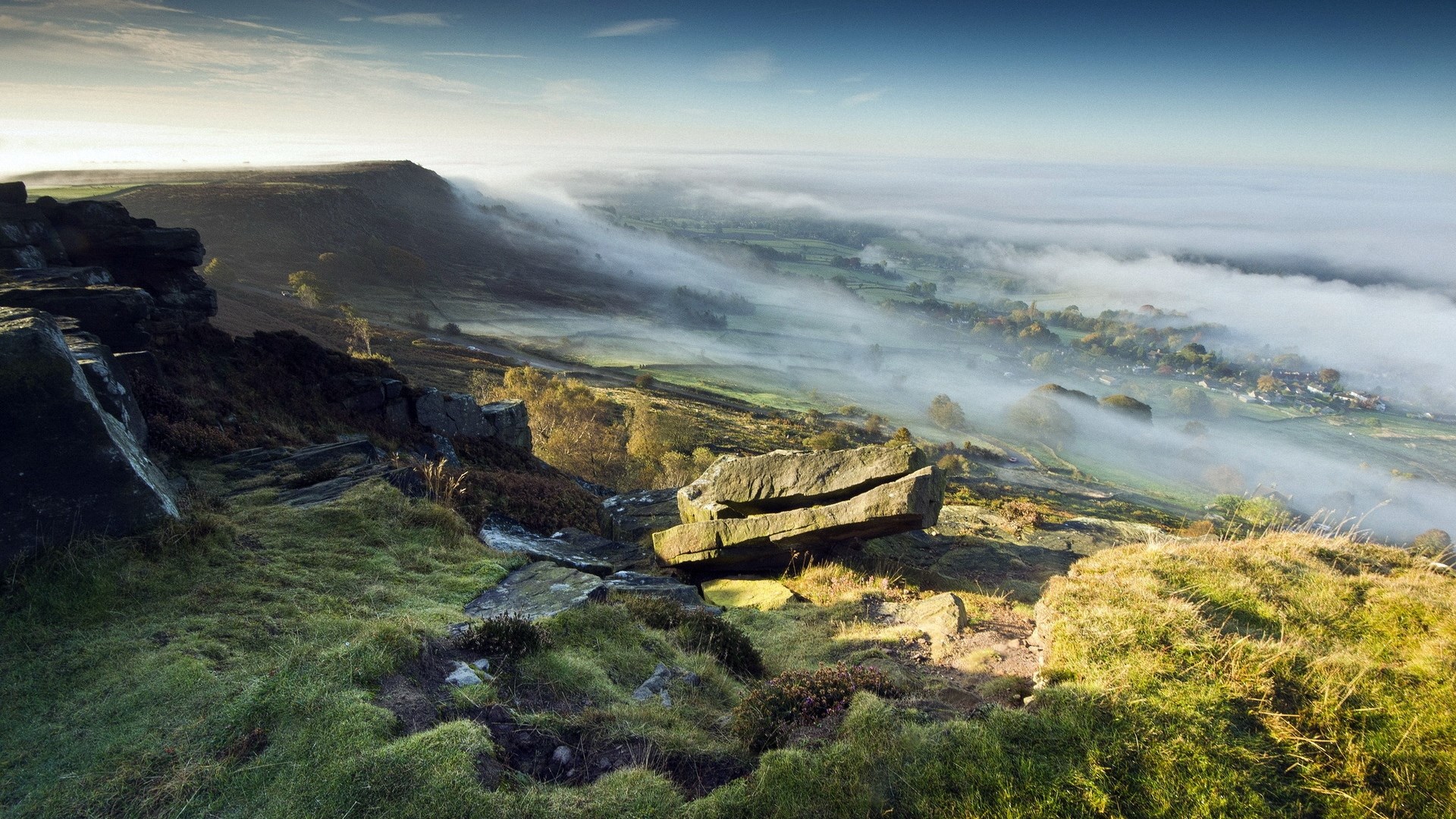 Misty valley with rocky foreground and distant village HD Wallpaper