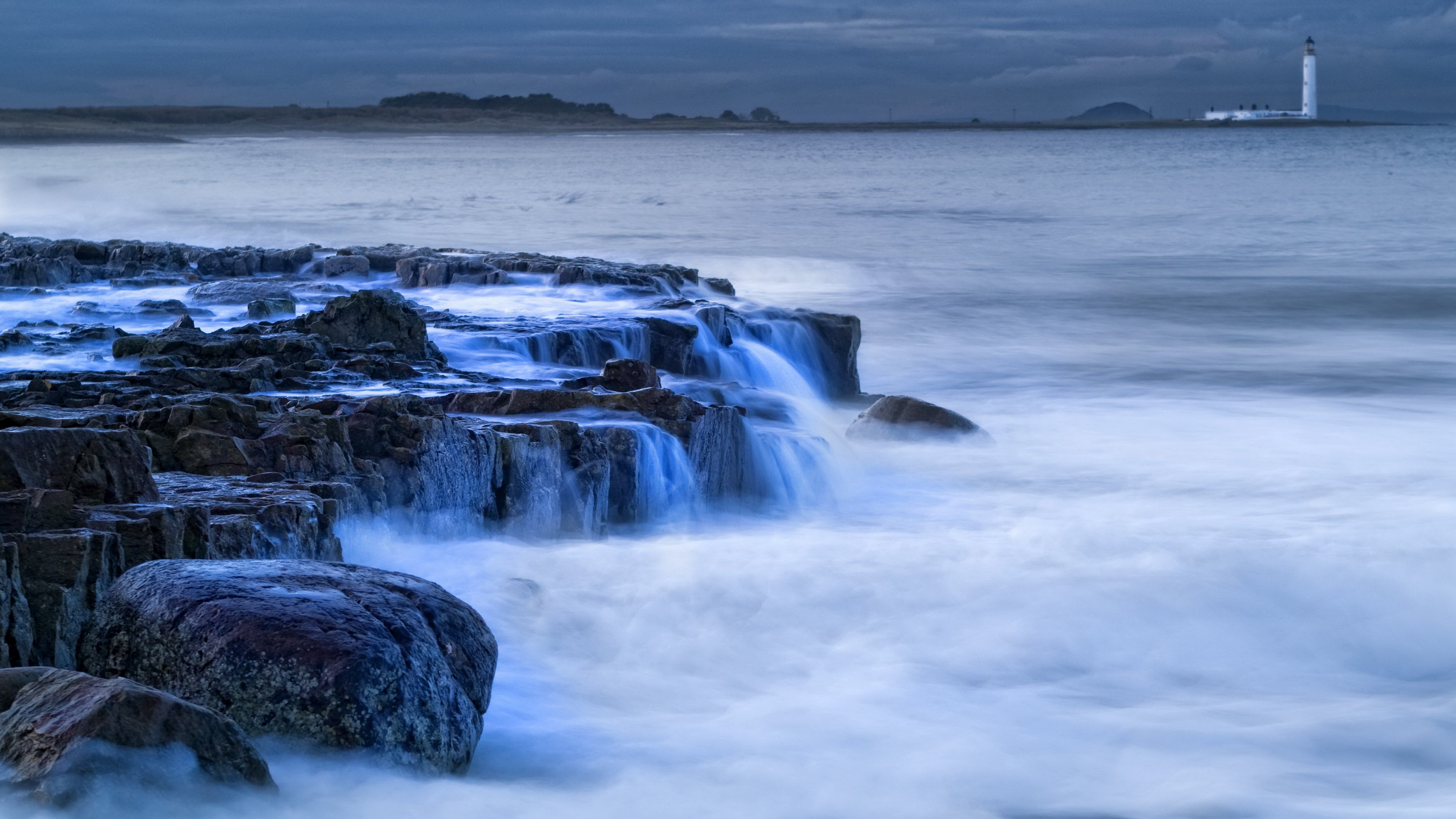 Coastal rocks with lighthouse in the distance HD Wallpaper