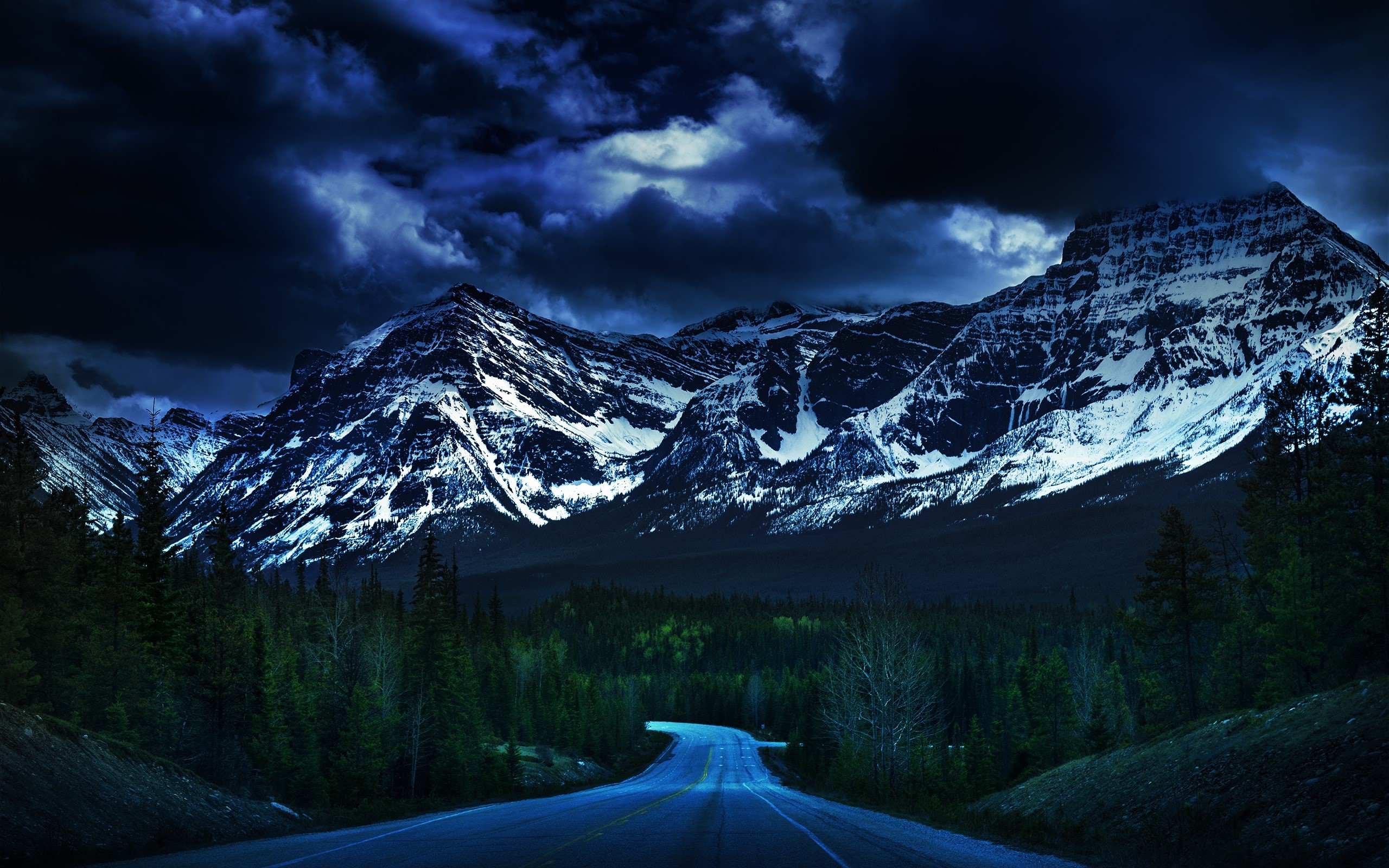 Road leading to snow-capped mountains under dark clouds HD Wallpaper