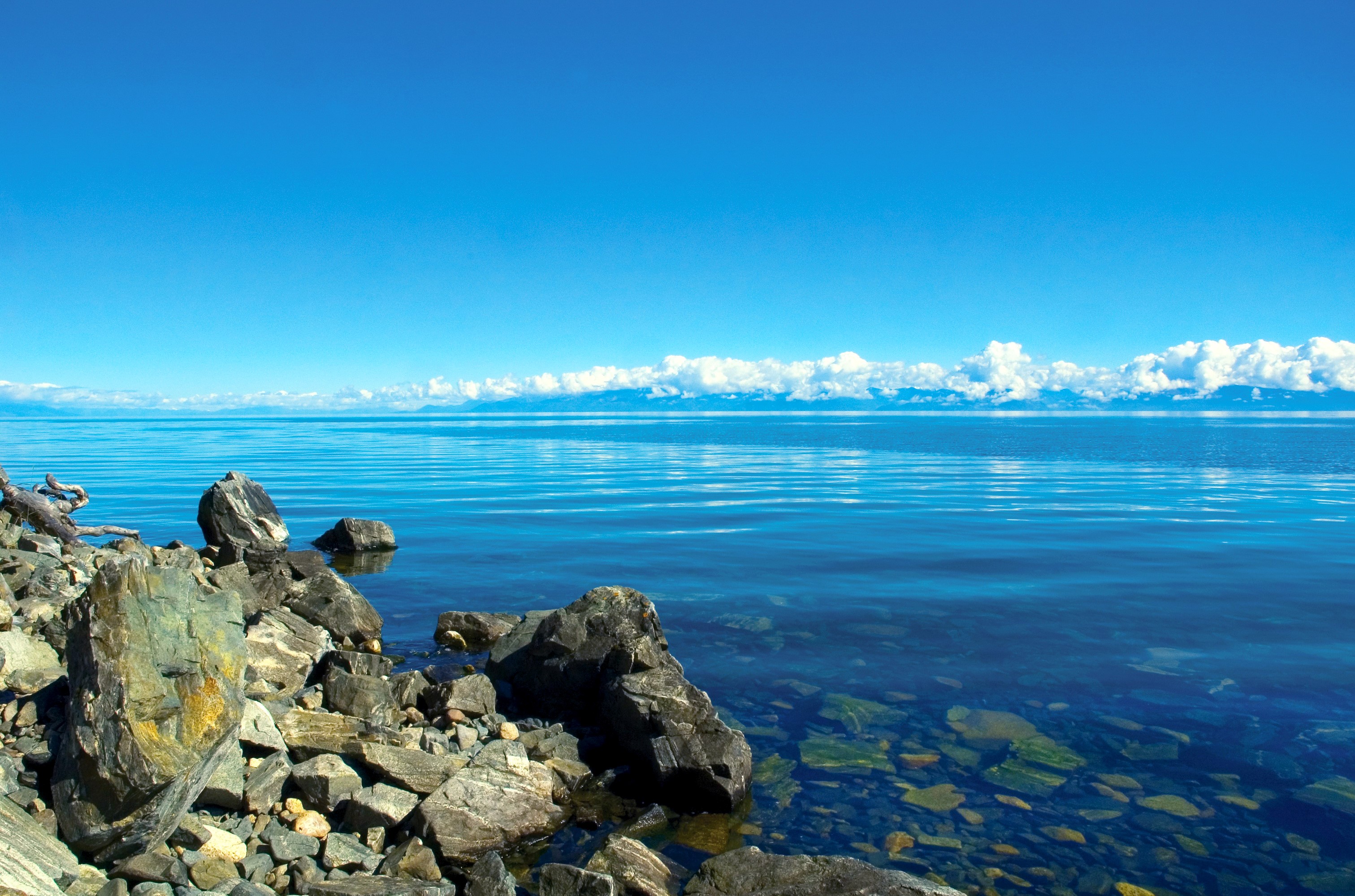 Rocky shore of a clear blue lake under a bright sky HD Wallpaper