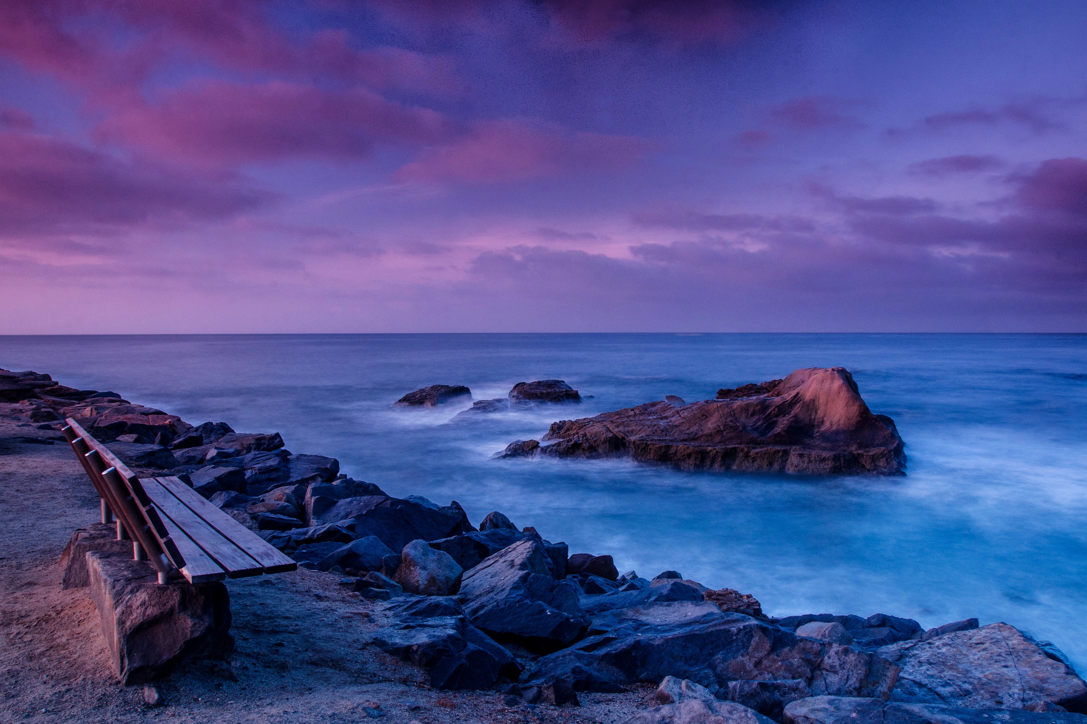 Bench on rocky coast with purple sky and ocean HD Wallpaper