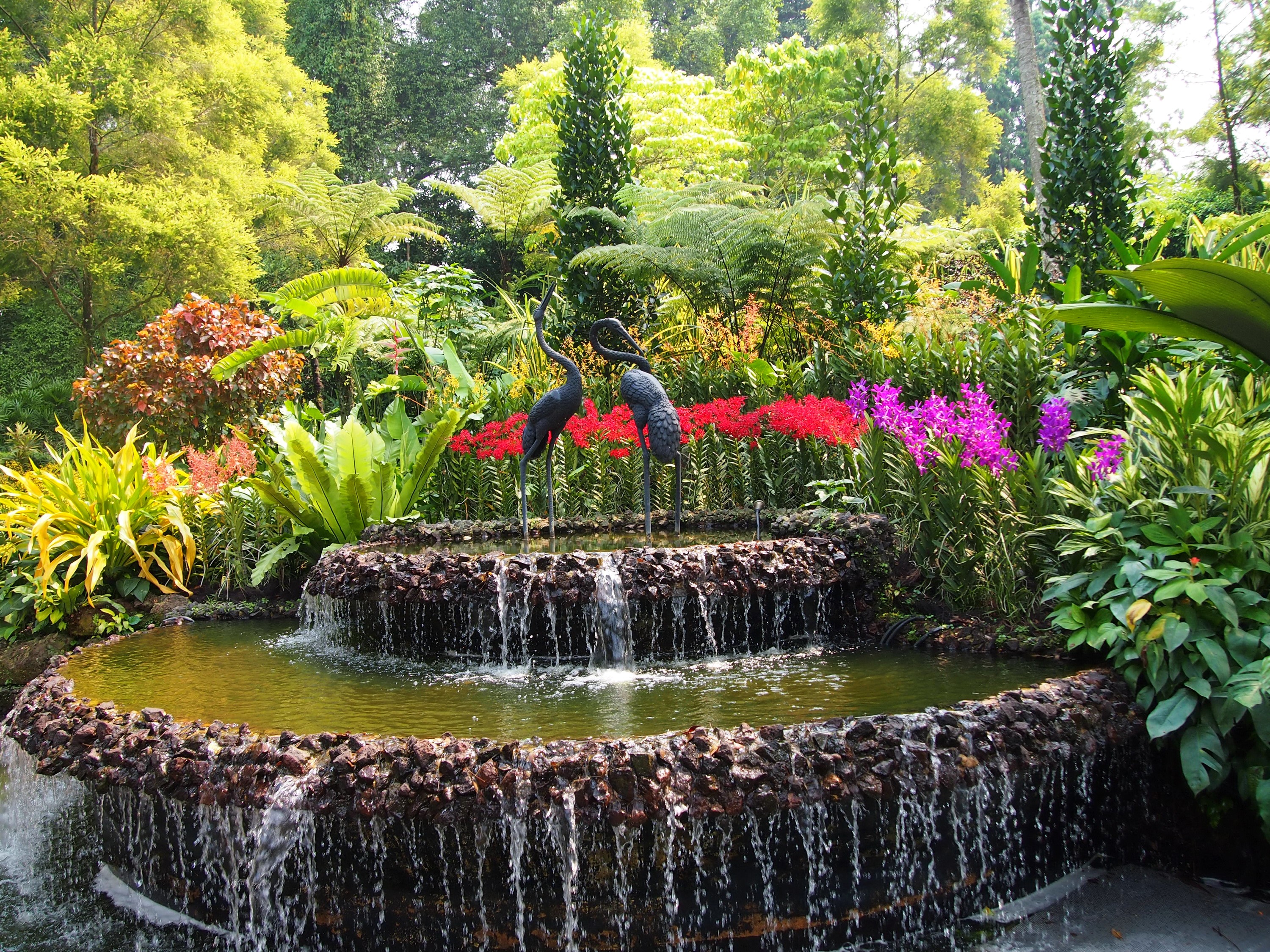 Tiered fountain with crane sculptures and colorful flowers in a garden HD Wallpaper