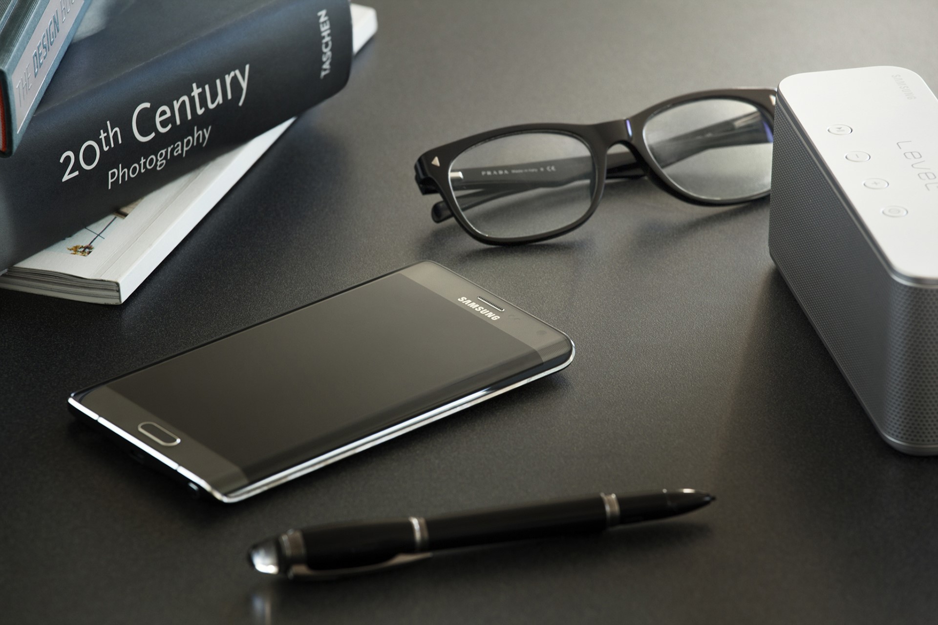 Samsung smartphone, glasses, pen, and speaker on desk HD Wallpaper