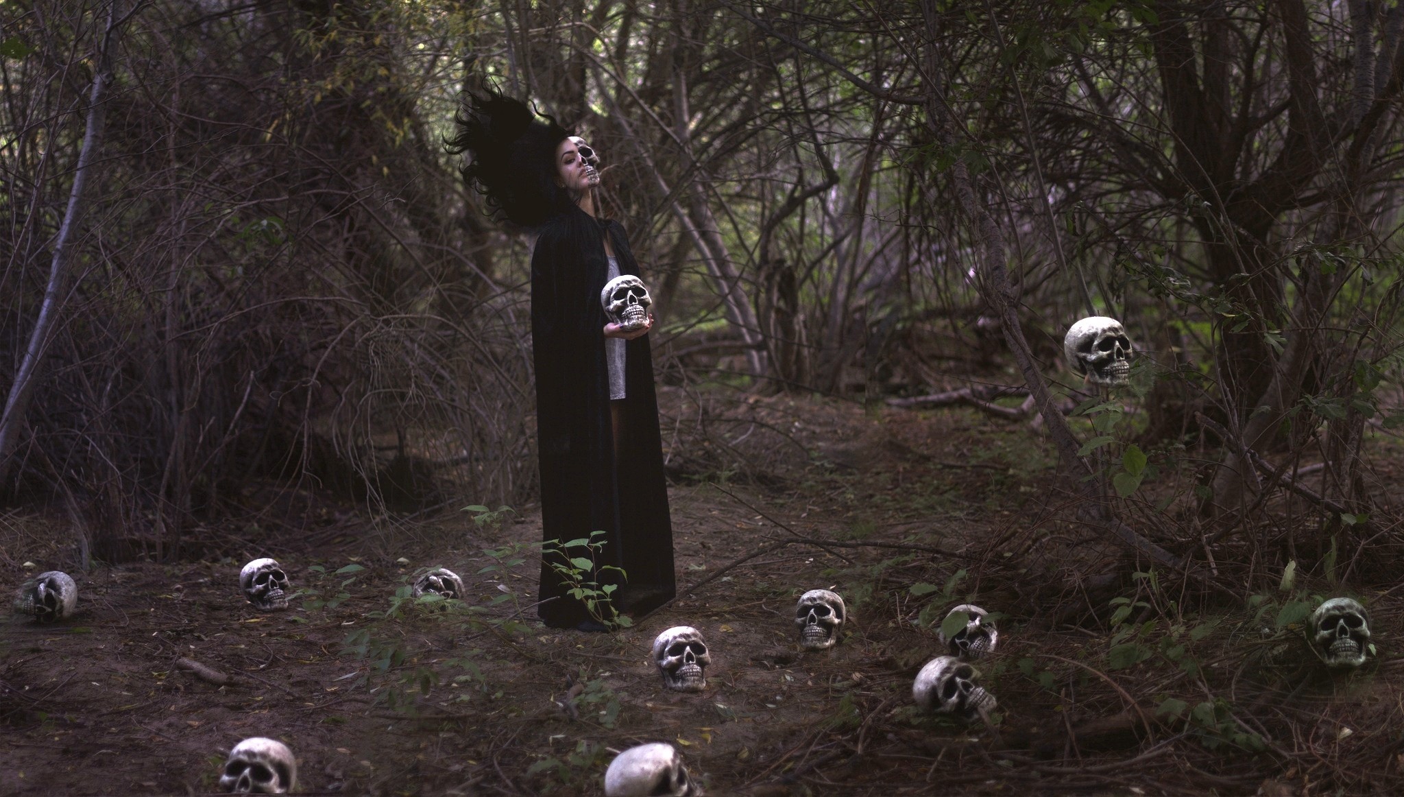 Woman in black cape holding a skull in a forest with skulls HD Wallpaper
