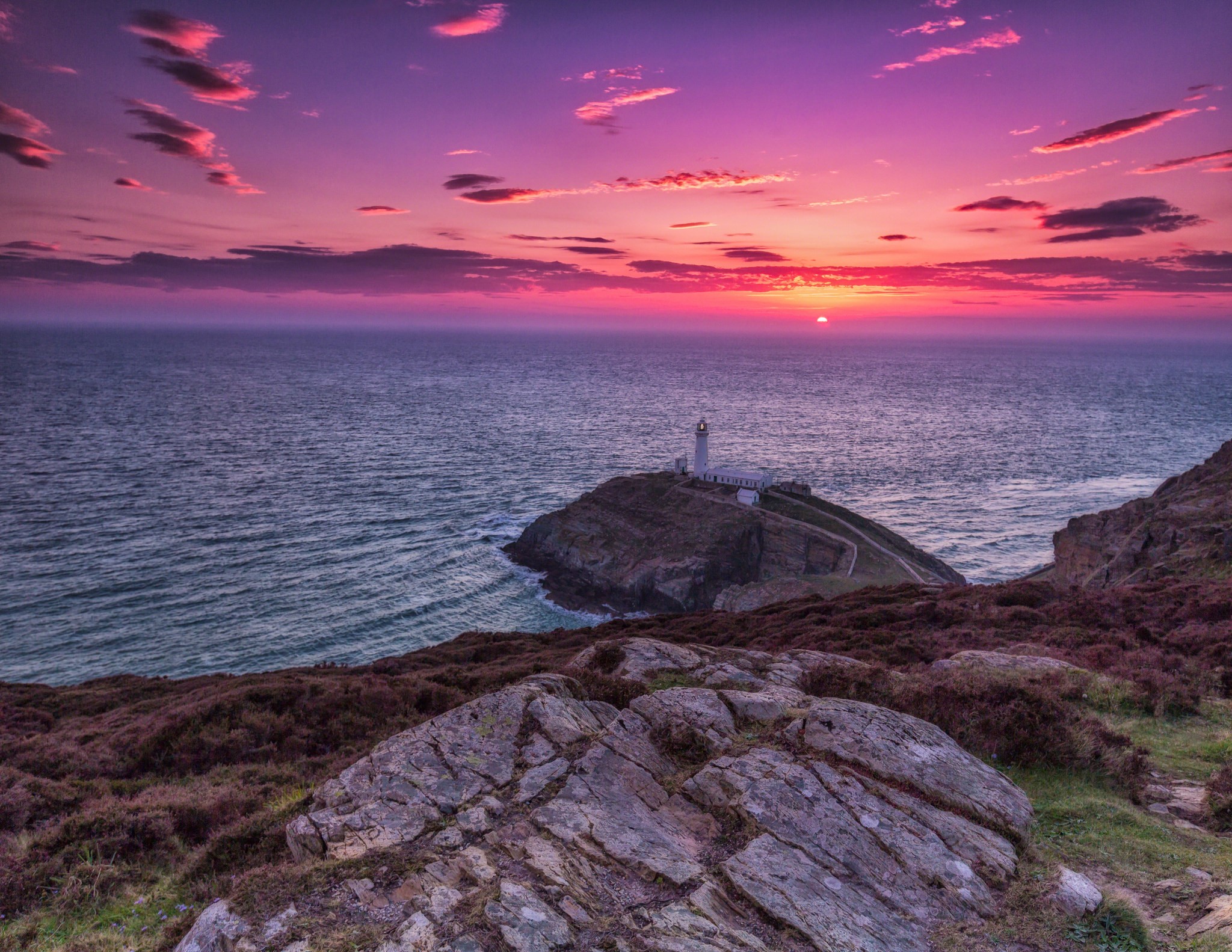 Lighthouse on a rocky outcrop during a vibrant sunset HD Wallpaper