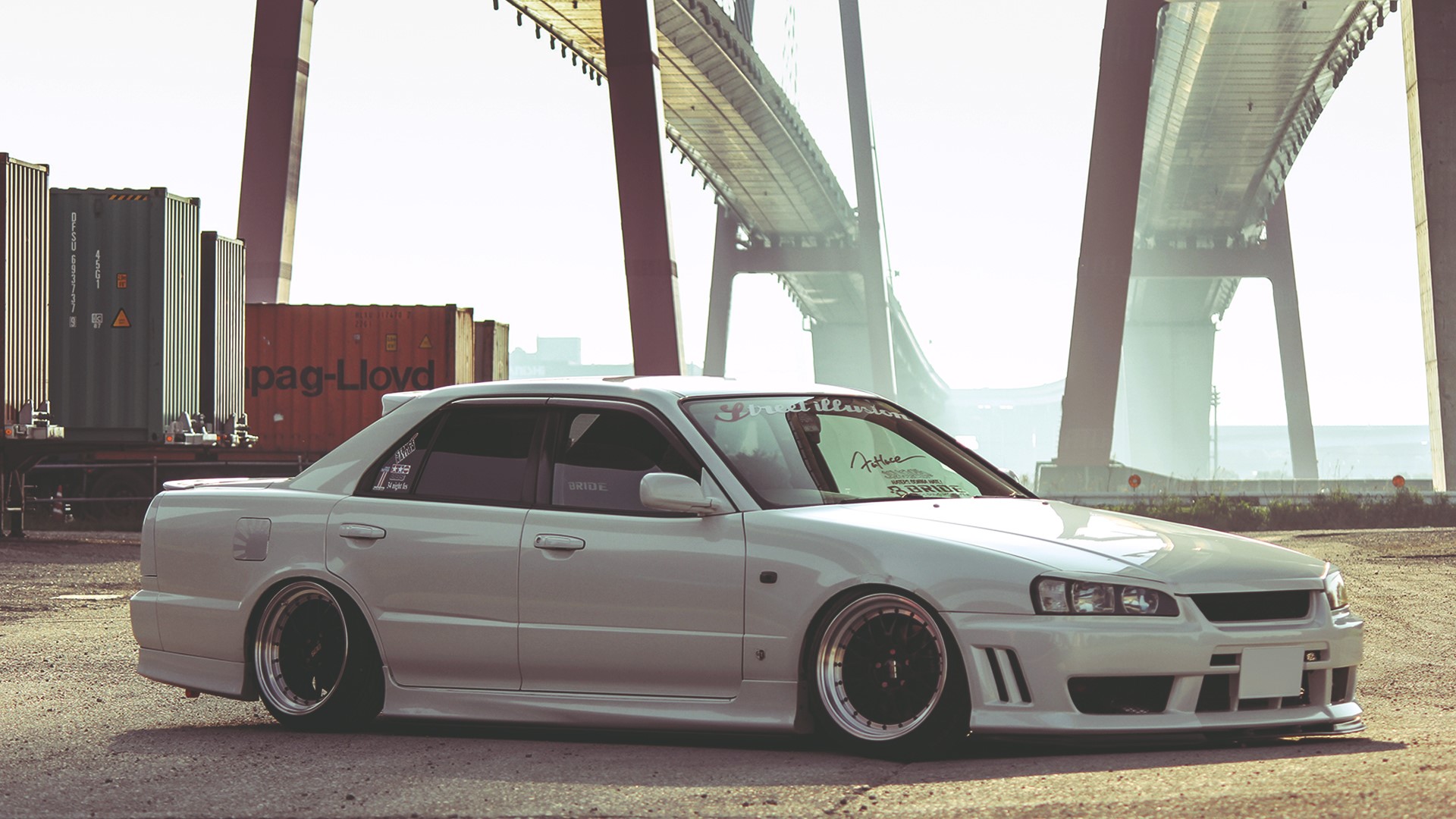 White Nissan Skyline sedan parked near shipping containers and a bridge HD Wallpaper