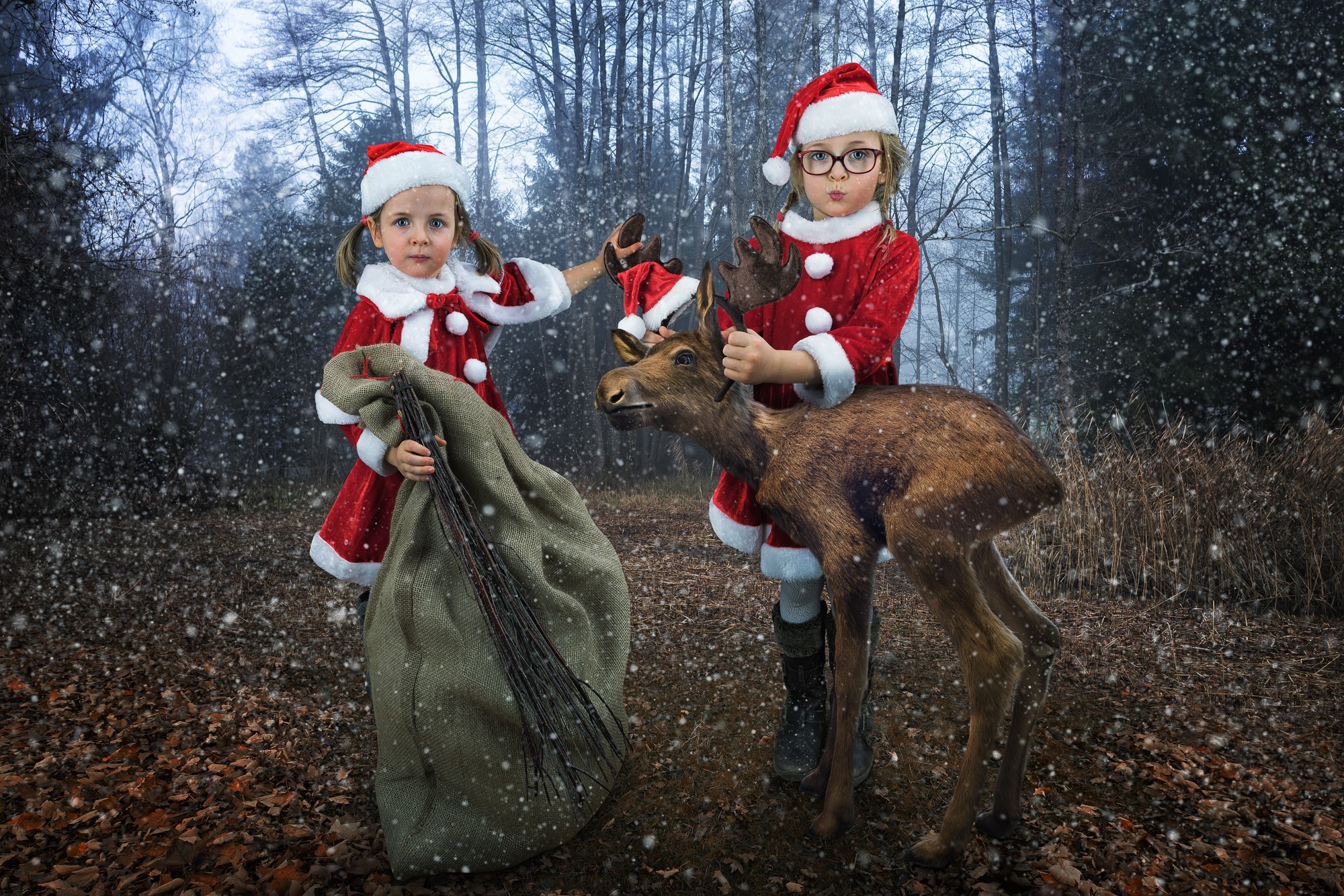 Two girls in Santa outfits with a deer in a snowy forest HD Wallpaper