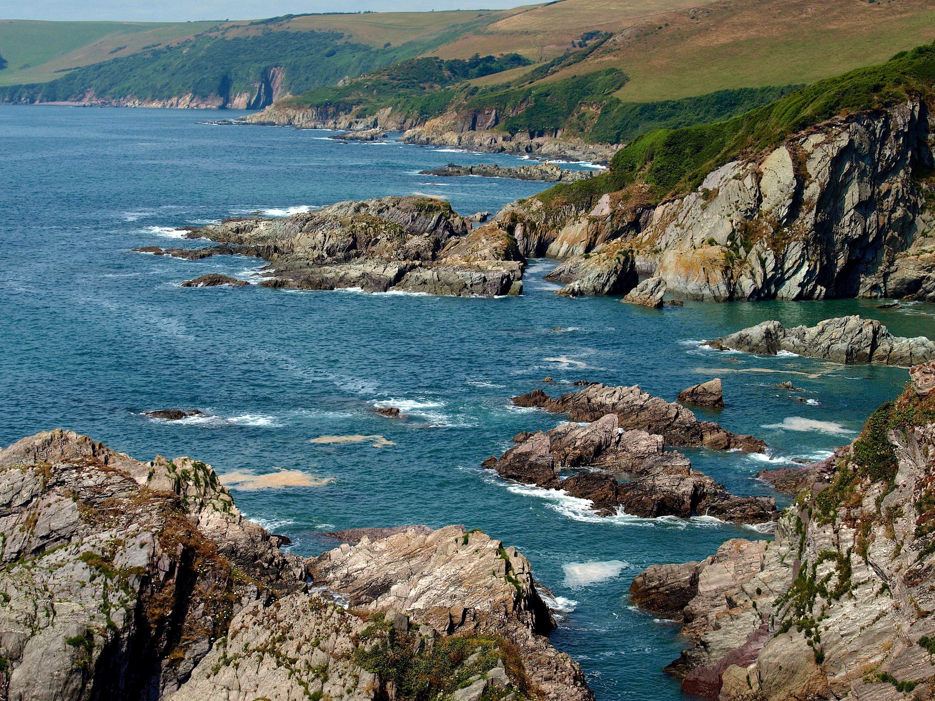 Rocky coastline with blue ocean and green hills HD Wallpaper