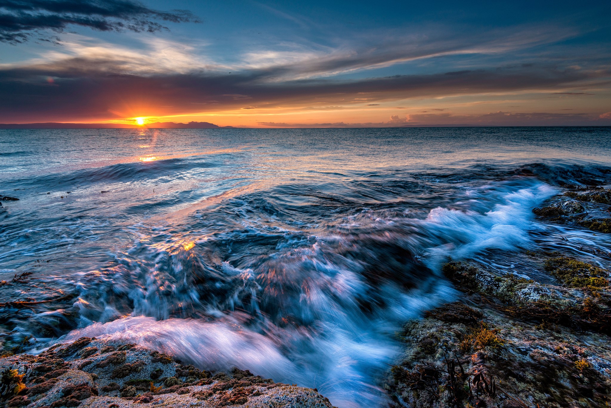 Sunset over the ocean with waves crashing on rocks HD Wallpaper