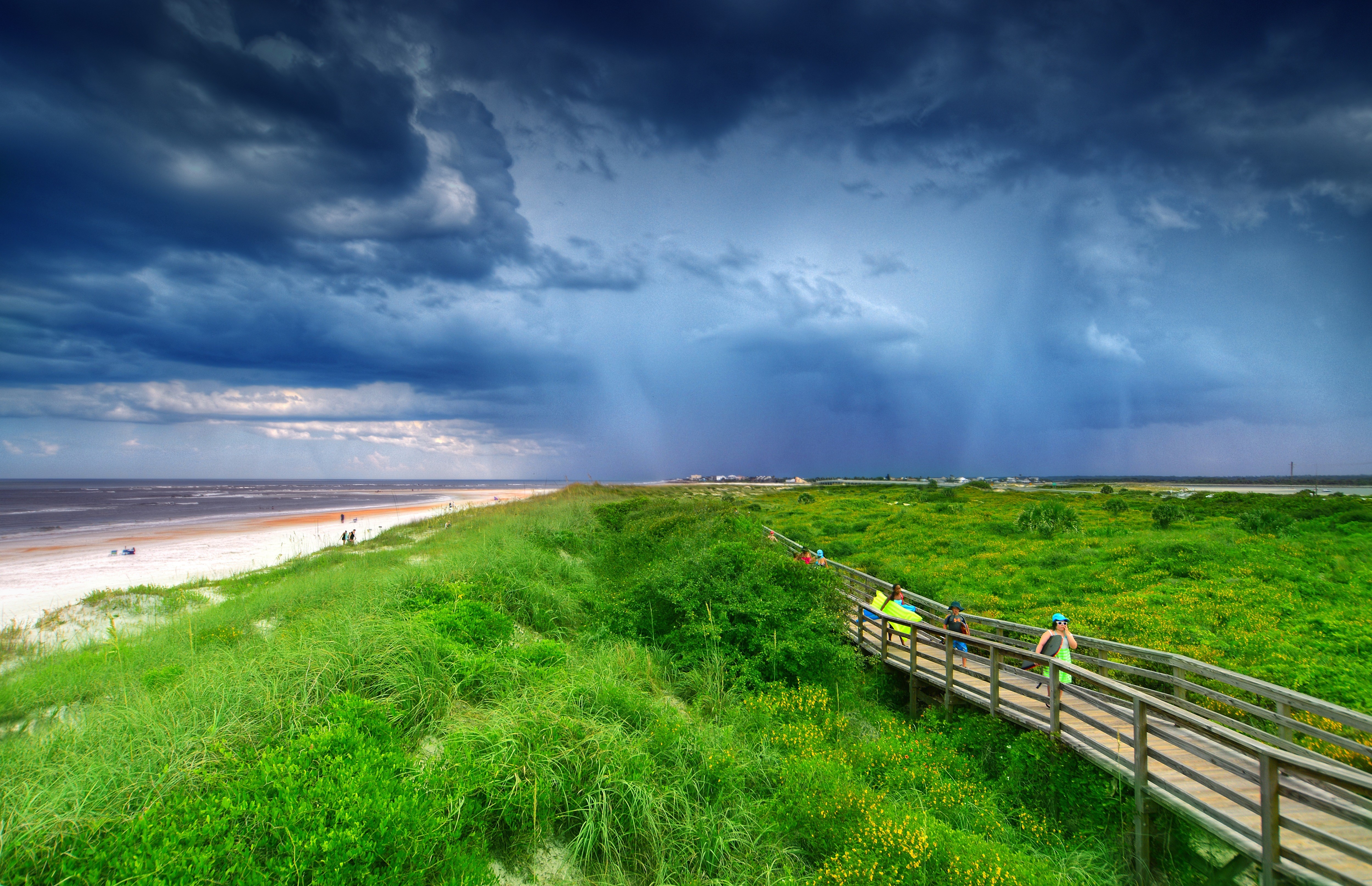 Wooden boardwalk through grassy dunes to a beach under stormy skies HD Wallpaper