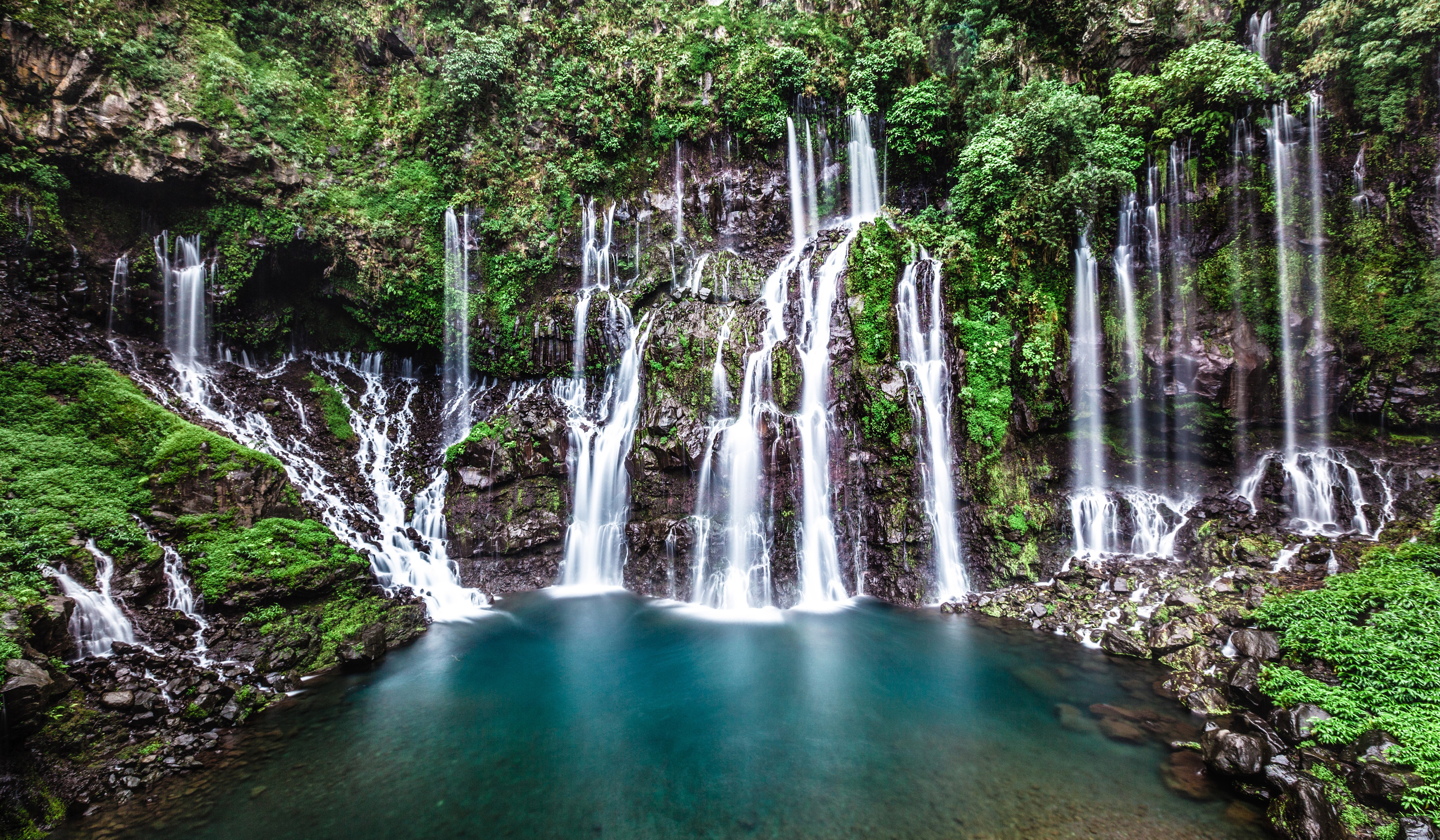 Multiple waterfalls cascade down mossy rocks into a turquoise pool HD Wallpaper