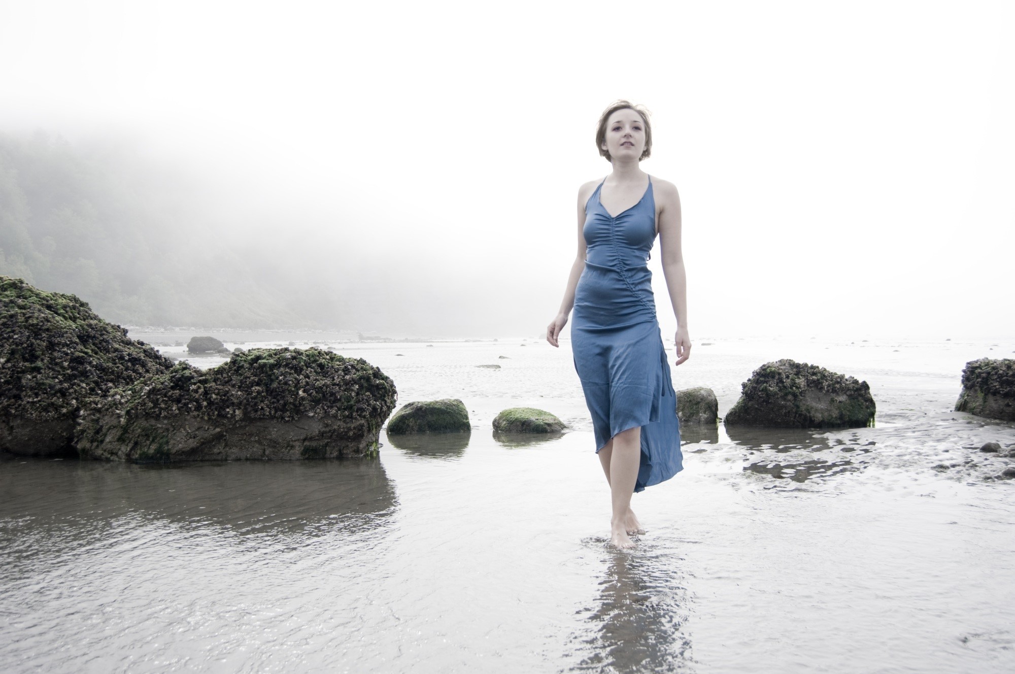 Woman in blue dress walking on a foggy beach with stones HD Wallpaper