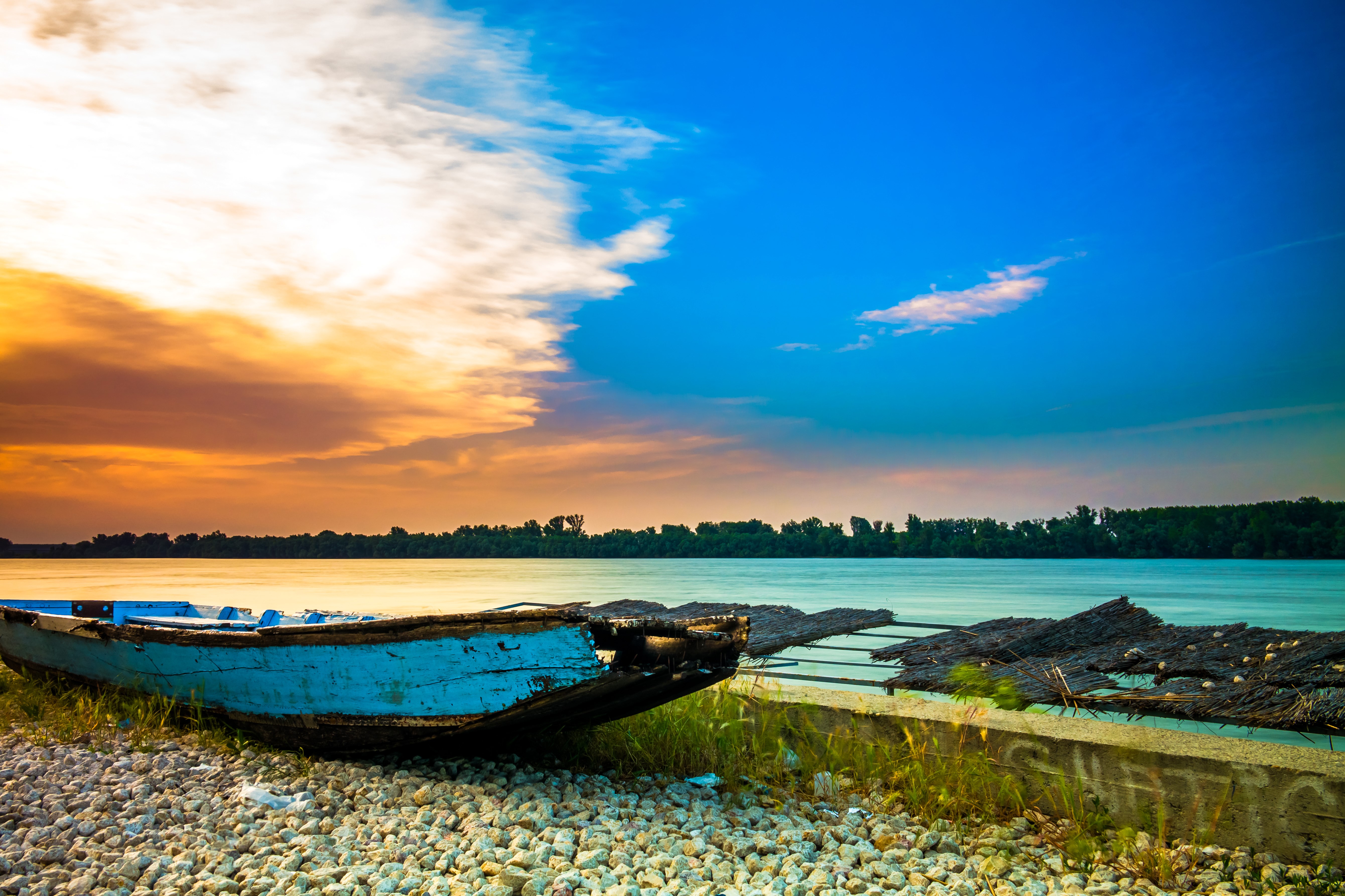 Old blue boat on a rocky shore by the river at sunset HD Wallpaper